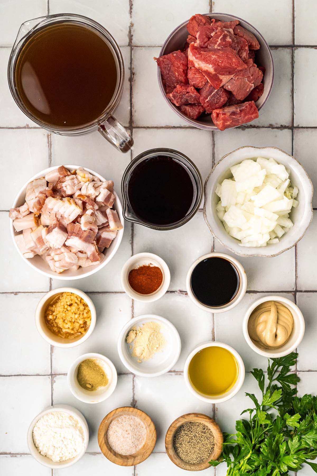 Bowls with beef, bacon, broth, sauce, chopped onion, seasonings, oil, and parsley on a white tiled surface.