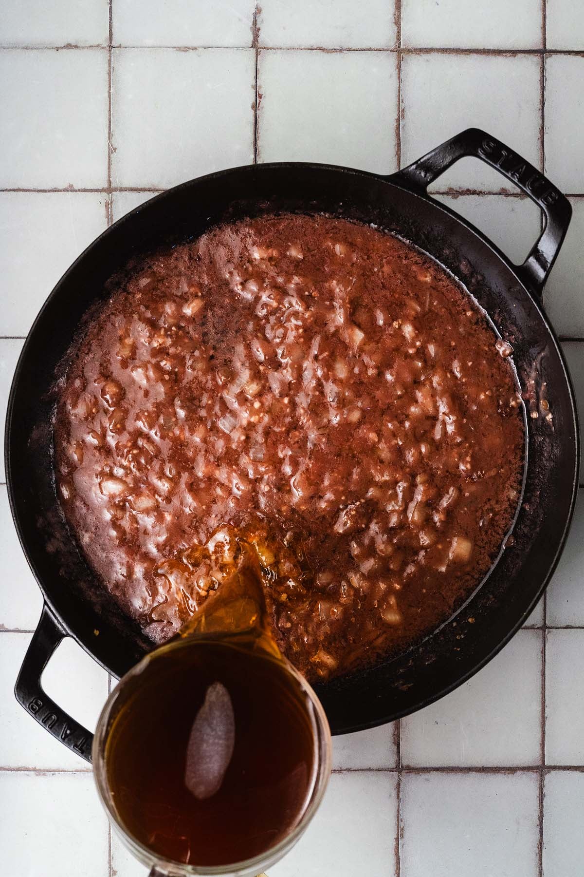 Broth is being poured from a measuring cup into a skillet with chopped onions and sauce on a tiled surface.