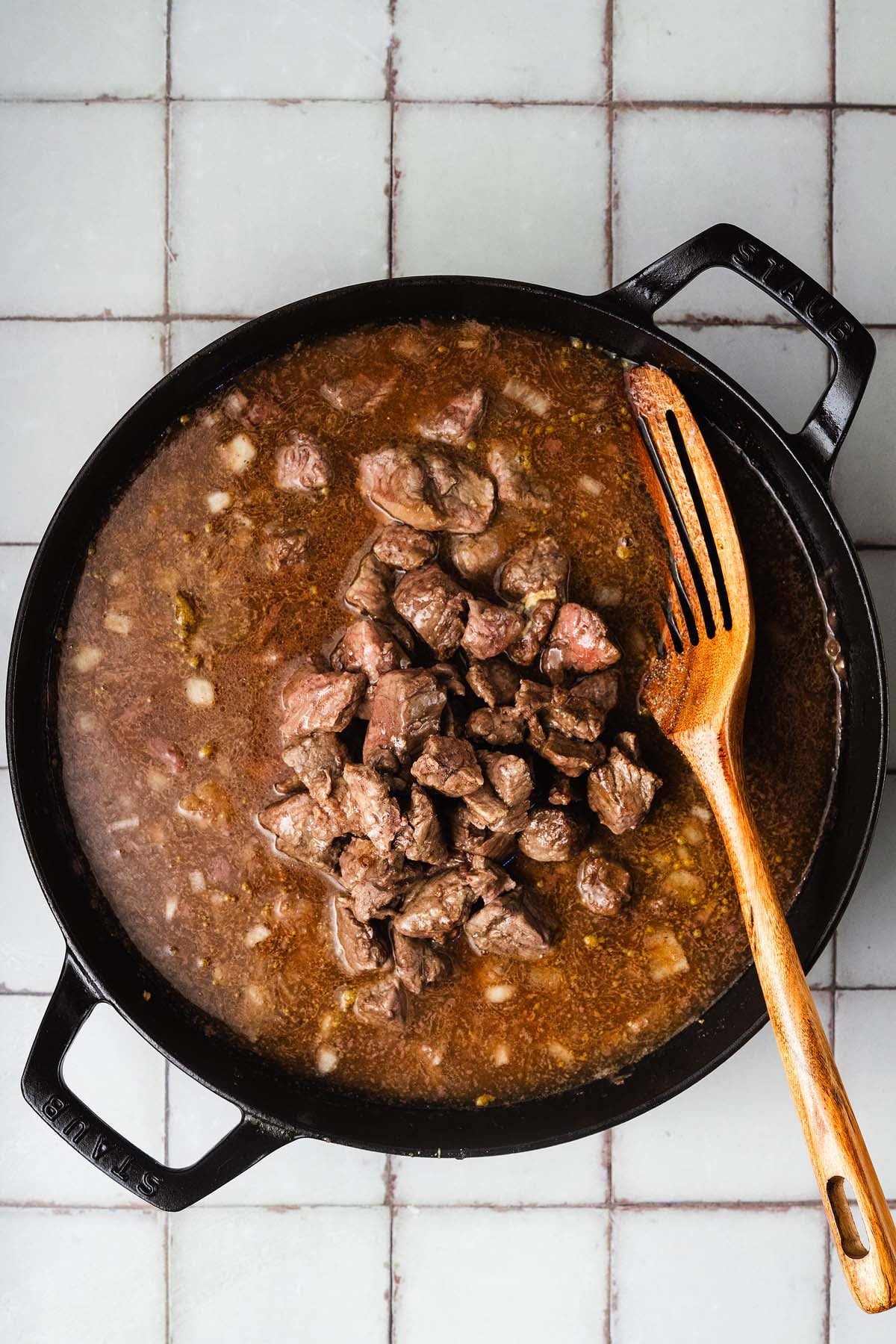 Chunks of beef simmer in broth in a black pot with a wooden spoon on a tiled countertop.