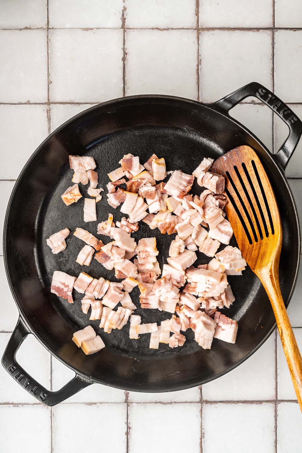 Chopped bacon cooking in a black skillet with a wooden spatula on a white tiled surface.