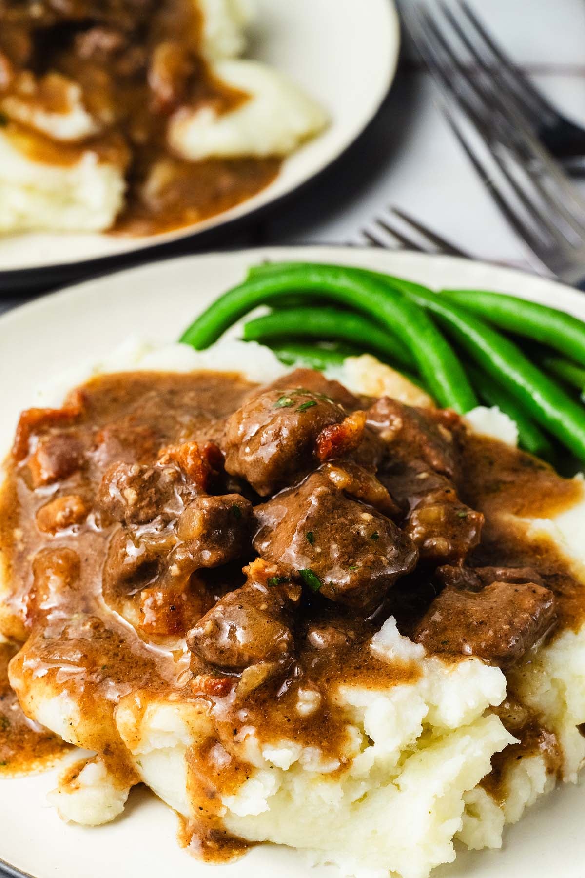 Beef chunks in a gravy over mashed potatoes with green beans on a plate, with a fork in the background.