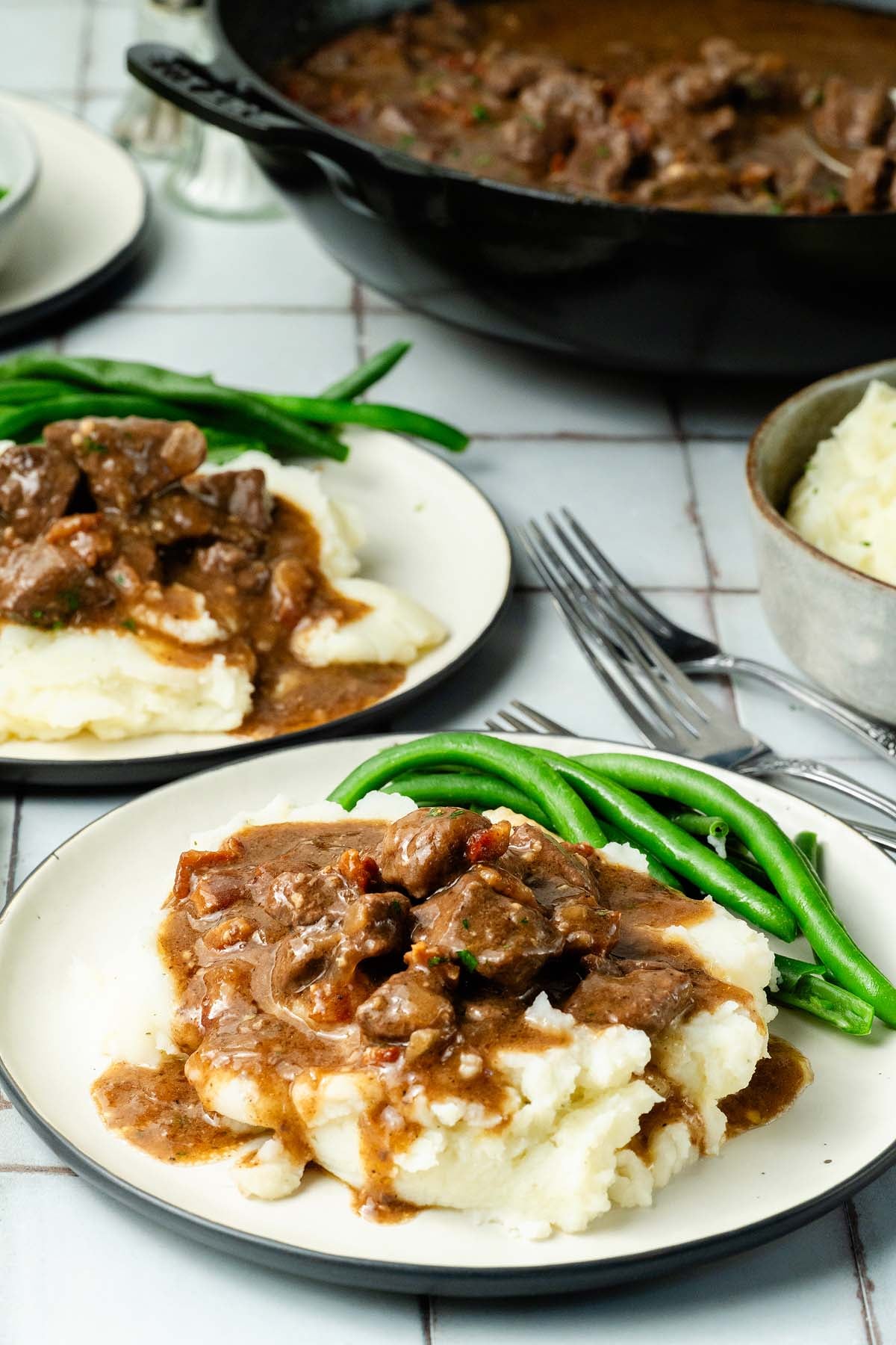 Gravy smothered beef tips over mashed potatoes with green beans on a plate, with more food in the background.