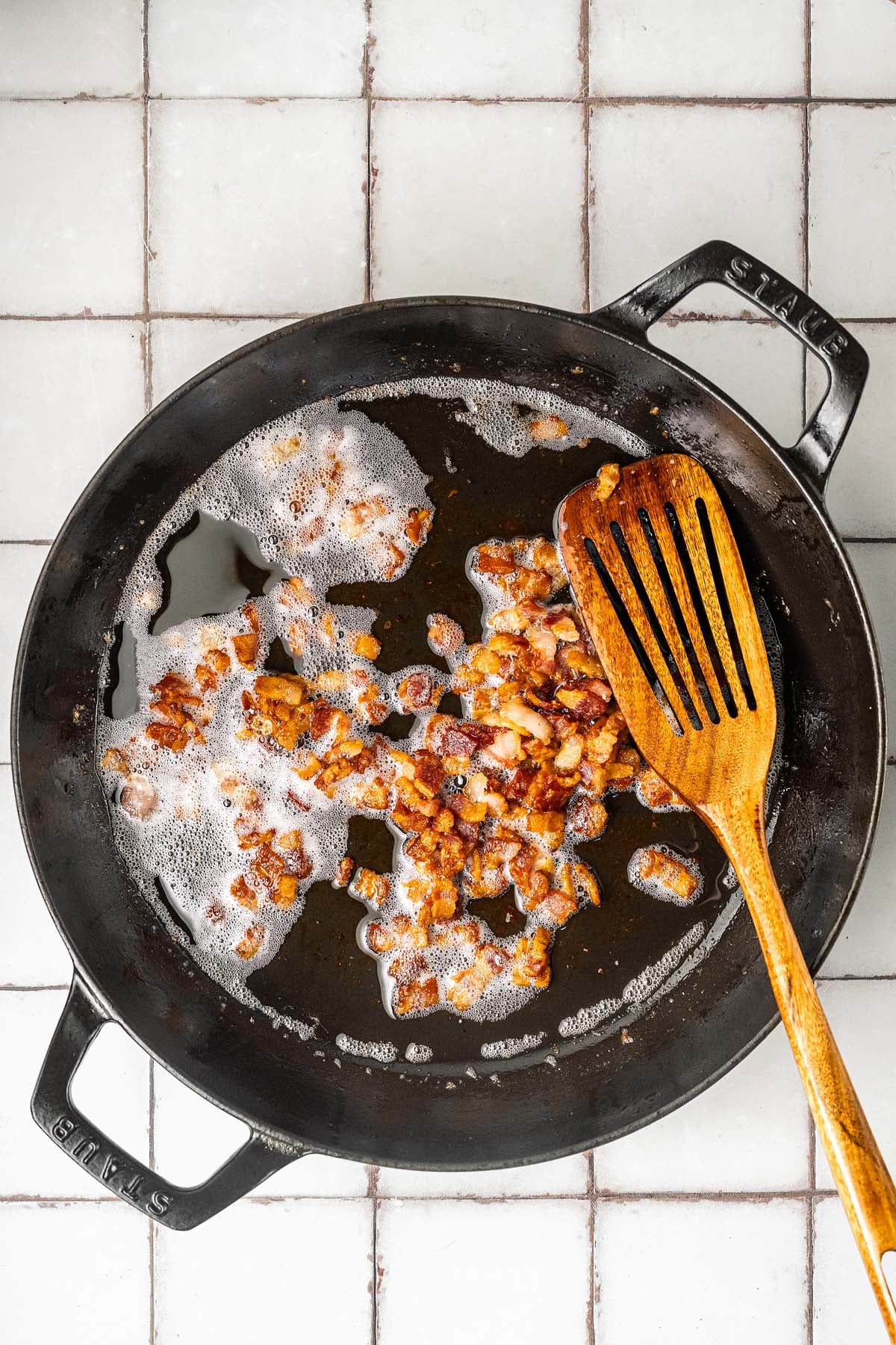 Bacon pieces sizzling in a black skillet with a wooden slotted spoon on a tiled surface.