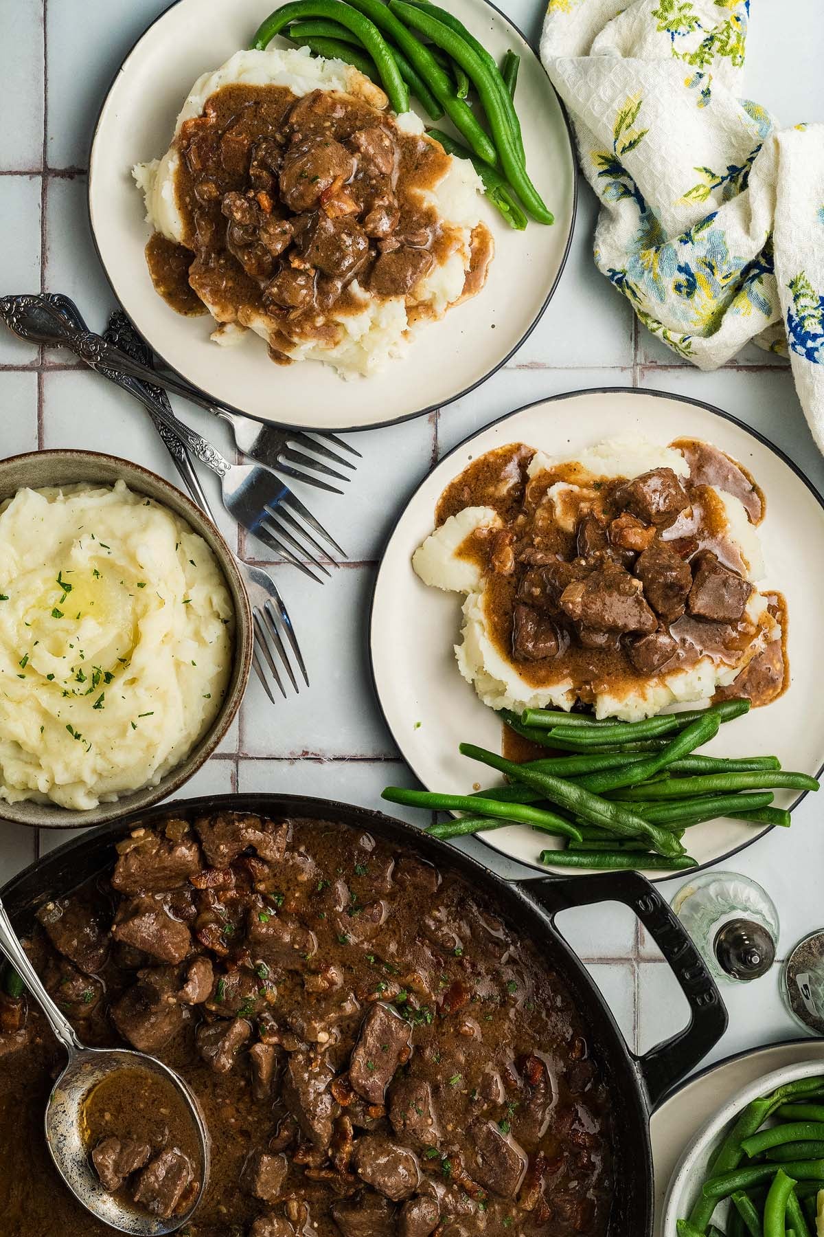 Two plates of beef tips in a gravy over mashed potatoes with green beans, plus a skillet and bowl of stew and potatoes.