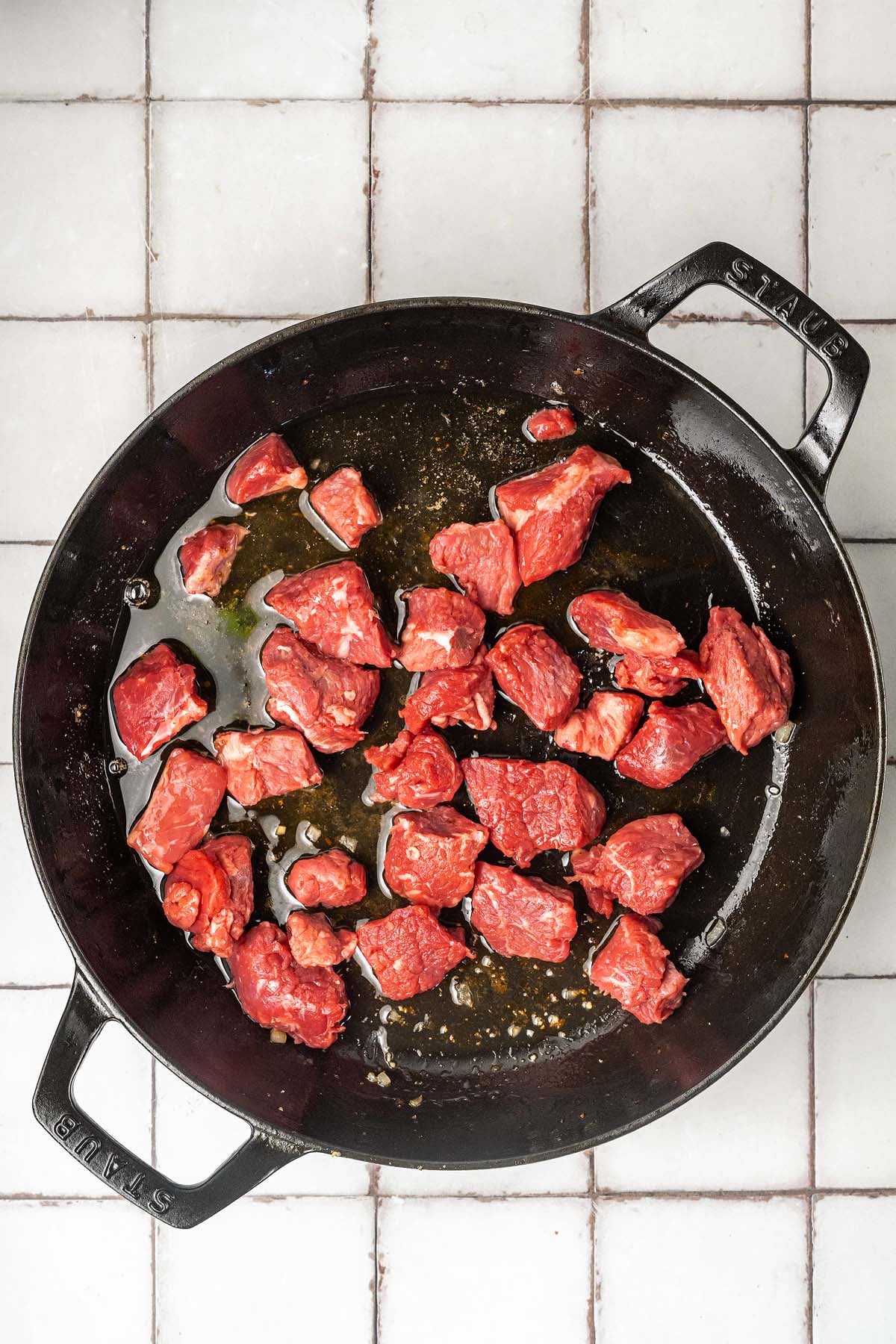 Chunks of raw beef in a black skillet with oil, on a white tiled countertop.