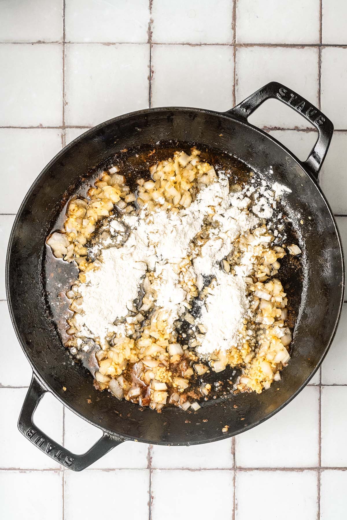Chopped onions and flour cooking in a black cast iron pan on a white tiled surface.