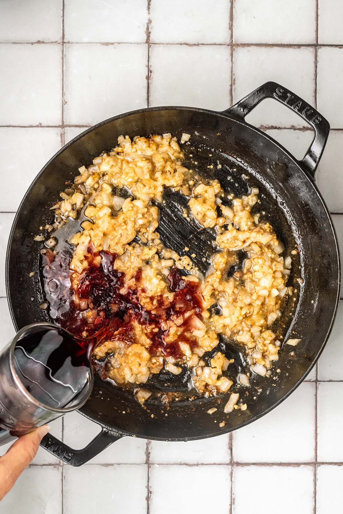 Hand pouring red wine into a skillet with sautéed onions and garlic on a tiled countertop.