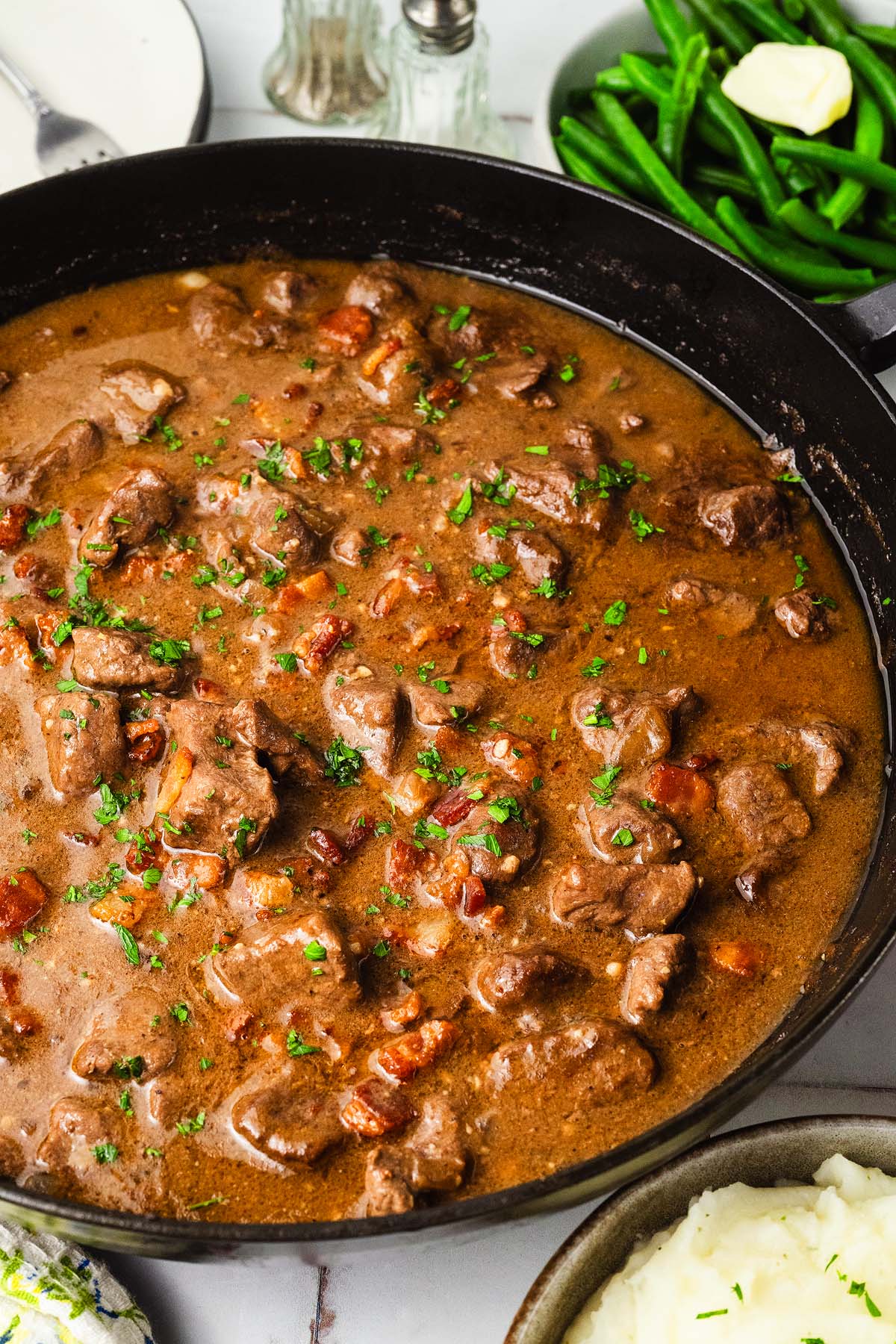 Smothered beef tips in a skillet, garnished with herbs. Green beans and mashed potatoes served on the side.