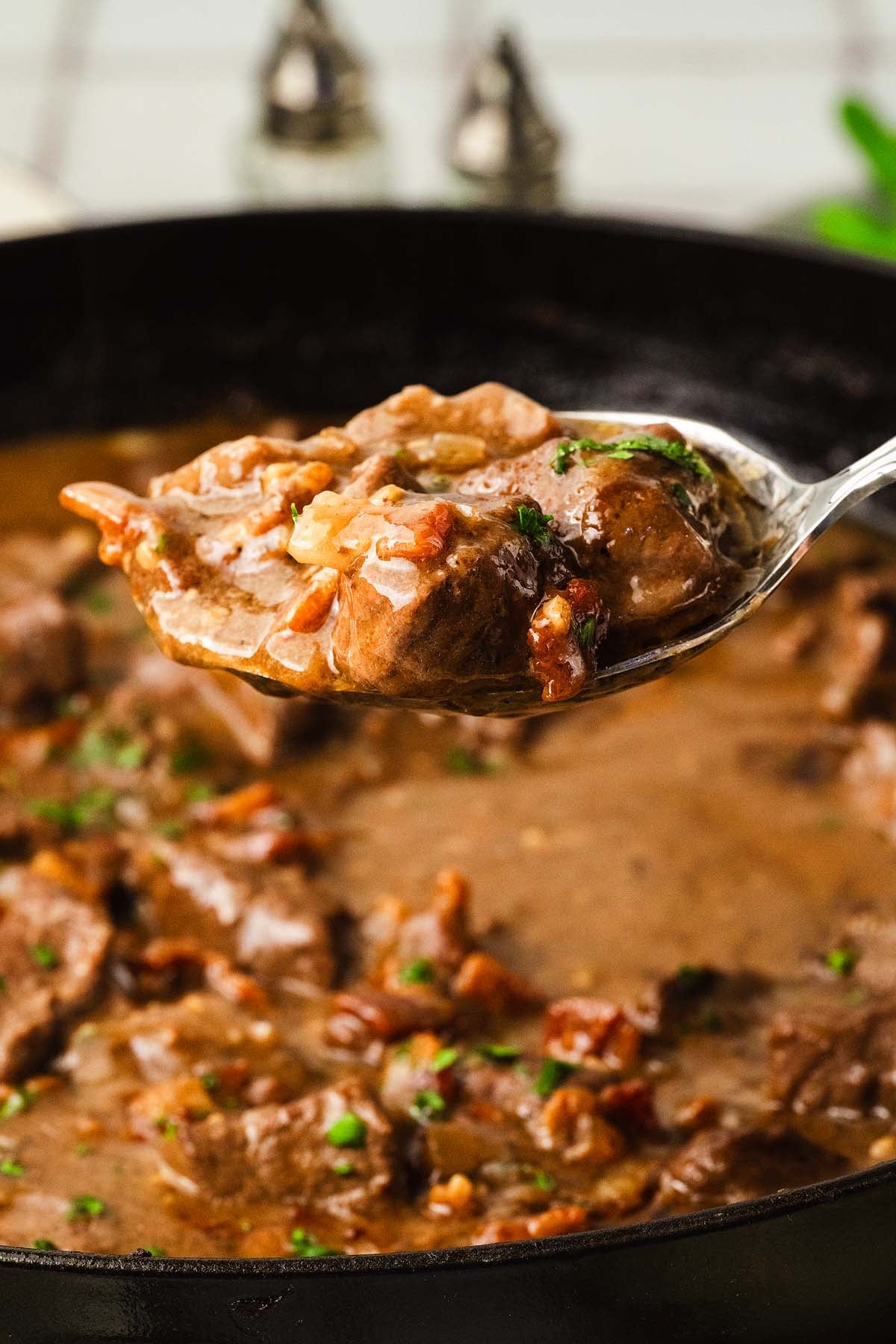 A spoonful of beef chunks with rich brown gravy, lifted above a pot, garnished with chopped herbs.