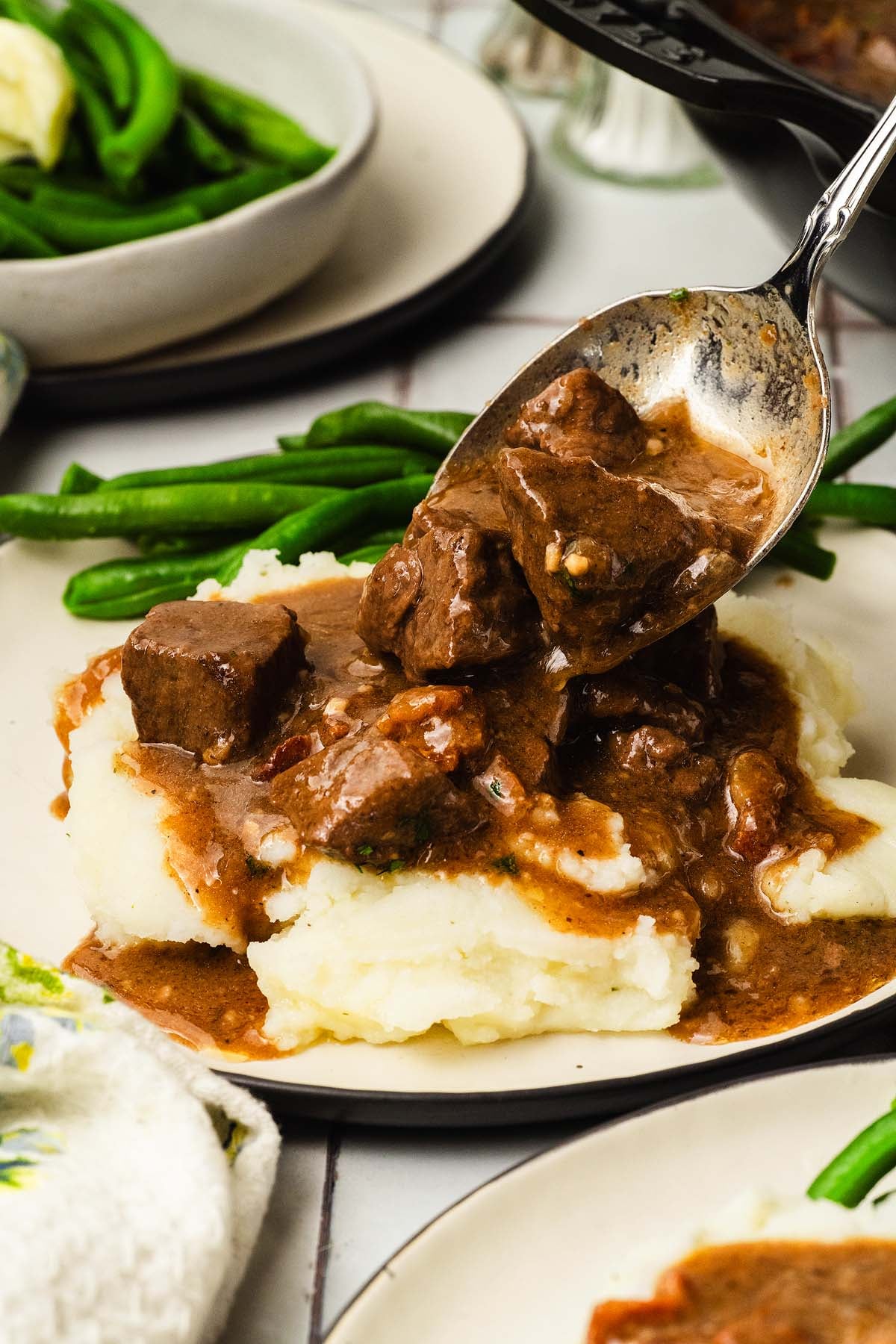A spoon serves beef tips in a gravy over mashed potatoes, with green beans on the side.