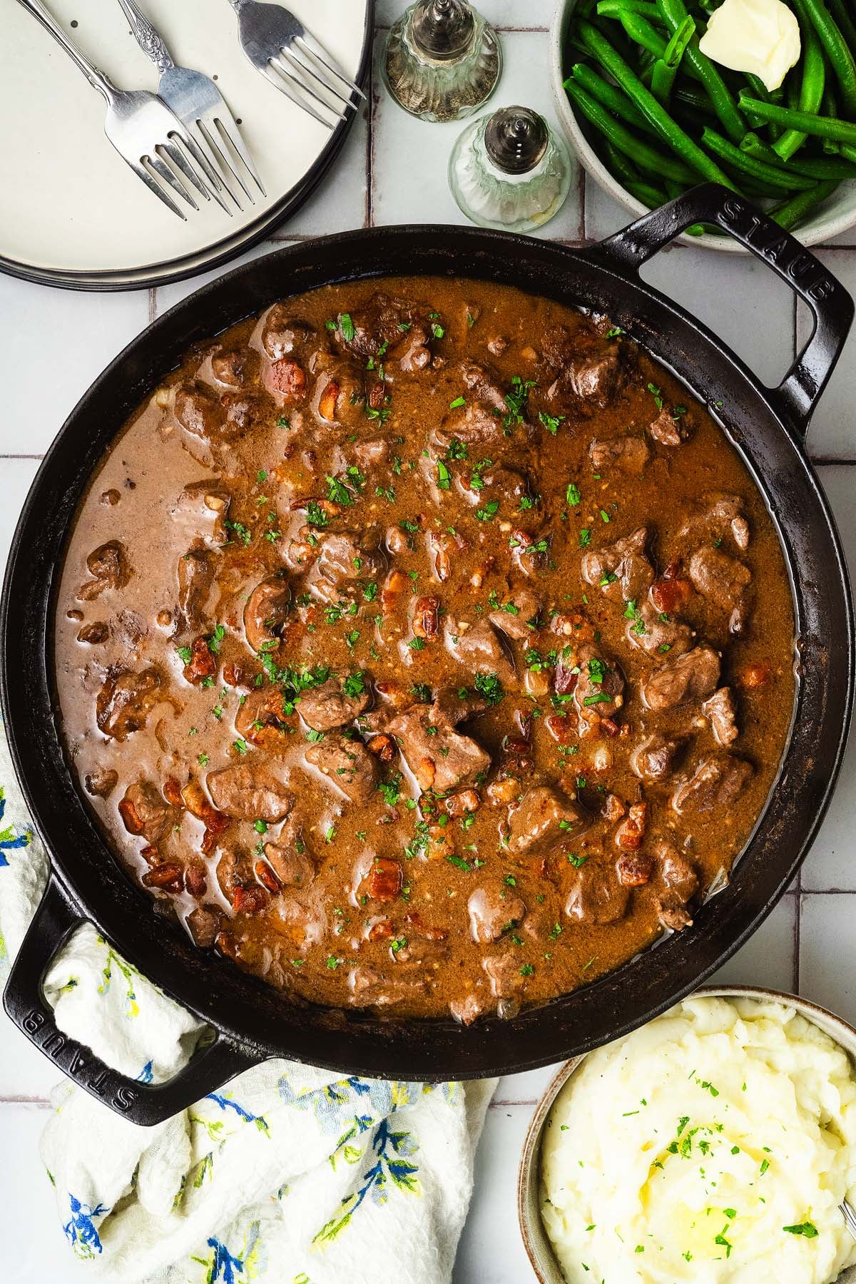 Gravy smothered beef tips in a skillet, served with mashed potatoes and green beans on the side.