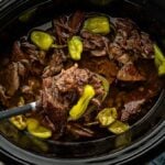 Shredded beef pot roast with pepperoncini in a slow cooker, a ladle lifting meat, with mashed potatoes on the side.