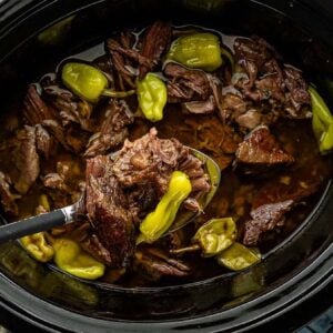 Shredded beef pot roast with pepperoncini in a slow cooker, a ladle lifting meat, with mashed potatoes on the side.