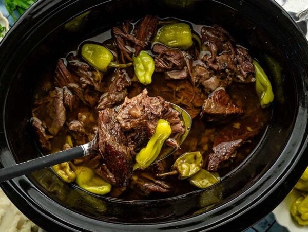 Shredded beef pot roast with pepperoncini in a slow cooker, a ladle lifting meat, with mashed potatoes on the side.