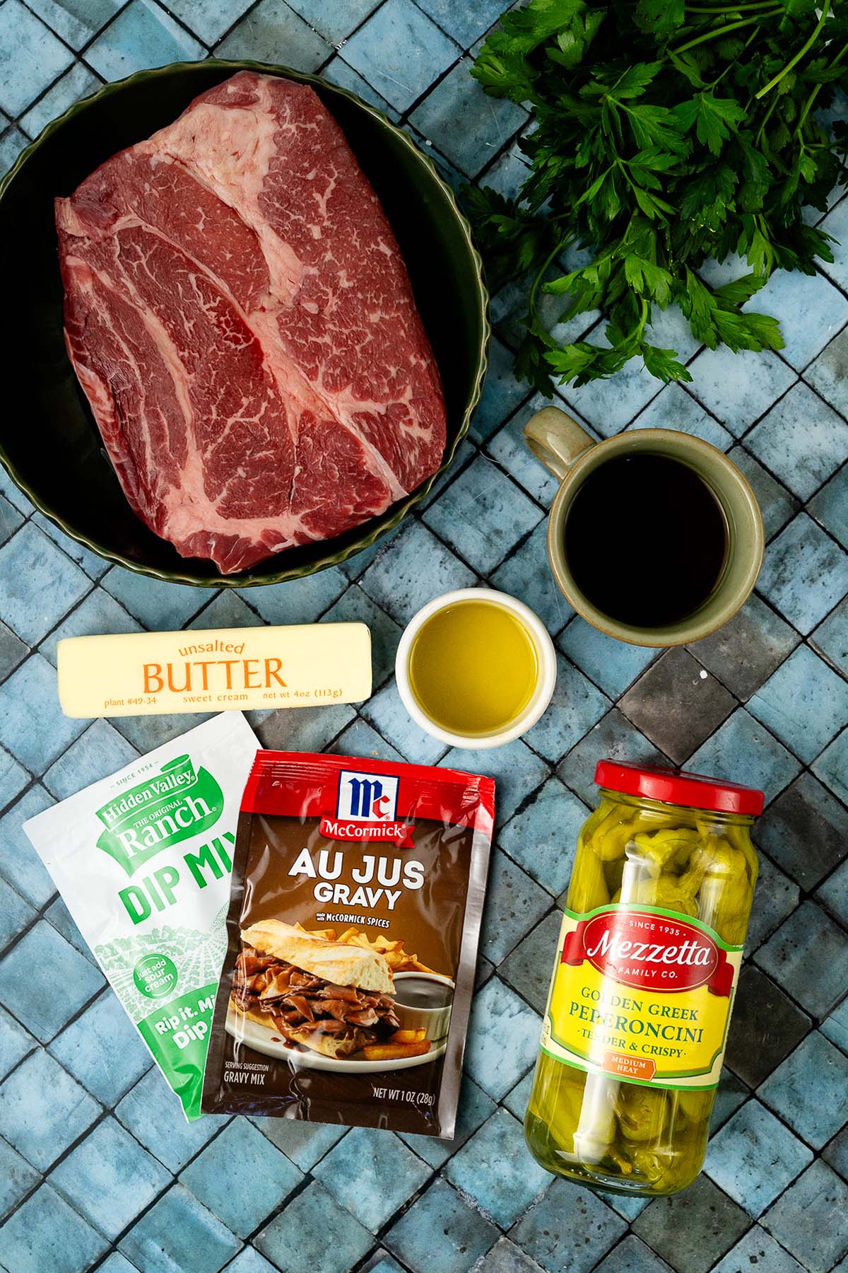 Raw beef, butter, ranch dip mix, au jus gravy, pepperoncini jar, coffee, oil, and parsley on blue tile.