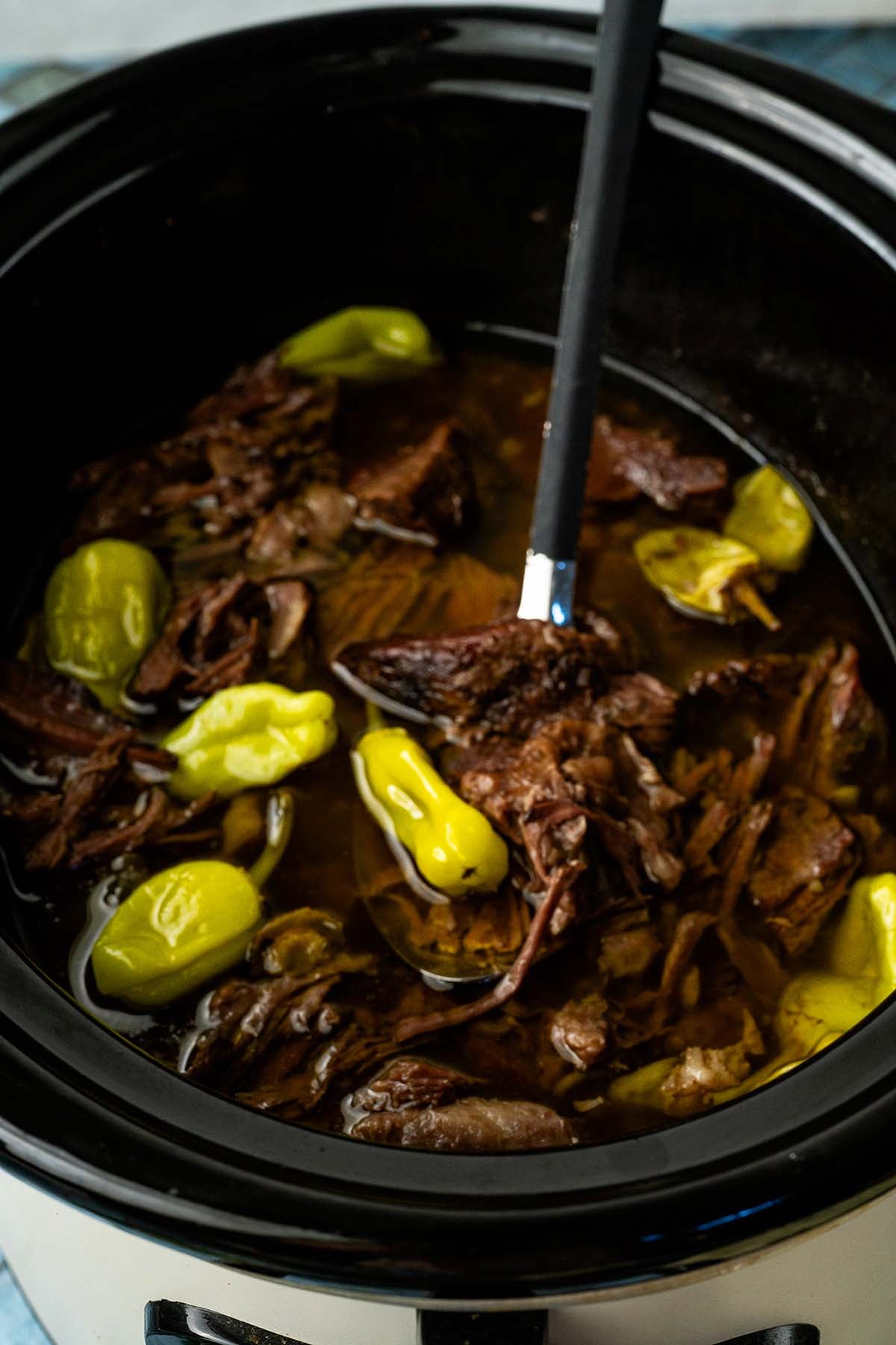Shredded beef and whole pepperoncini peppers simmering in a slow cooker, with a ladle inside.