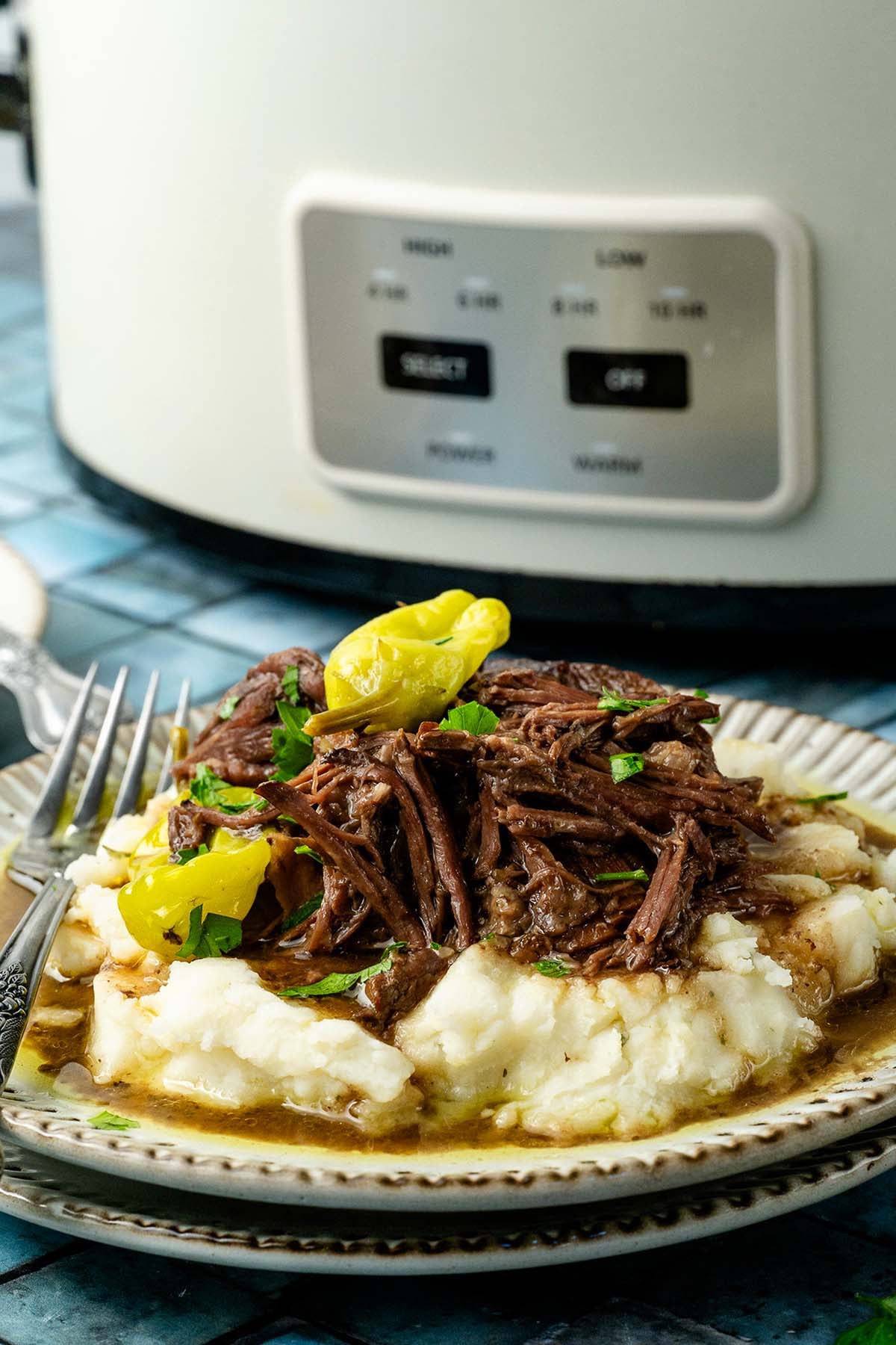 Plate of mashed potatoes topped with shredded roast beef, gravy, and pepperoncini, with a fork beside, slow cooker behind.