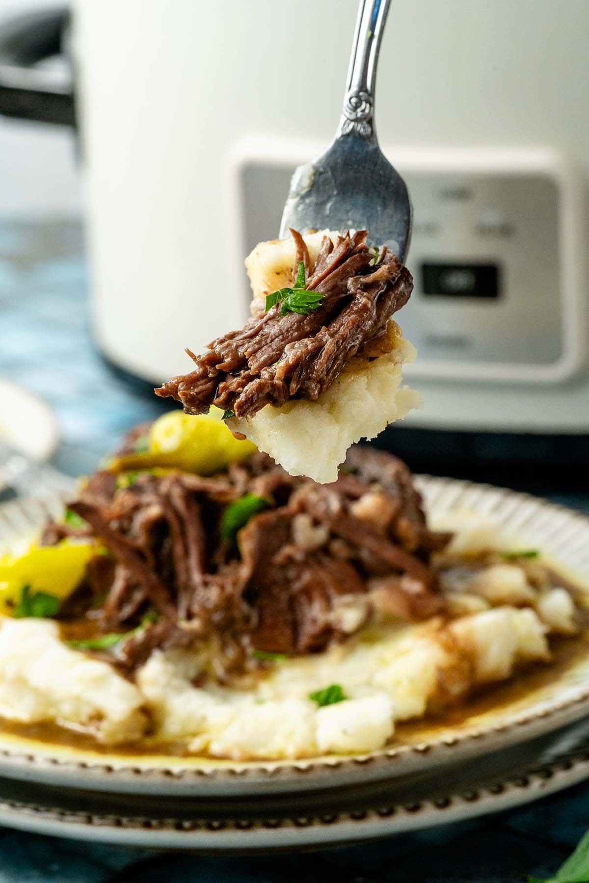 A fork holds shredded roasted beef and mashed potatoes above a plate with more beef and potatoes, garnished with herbs.