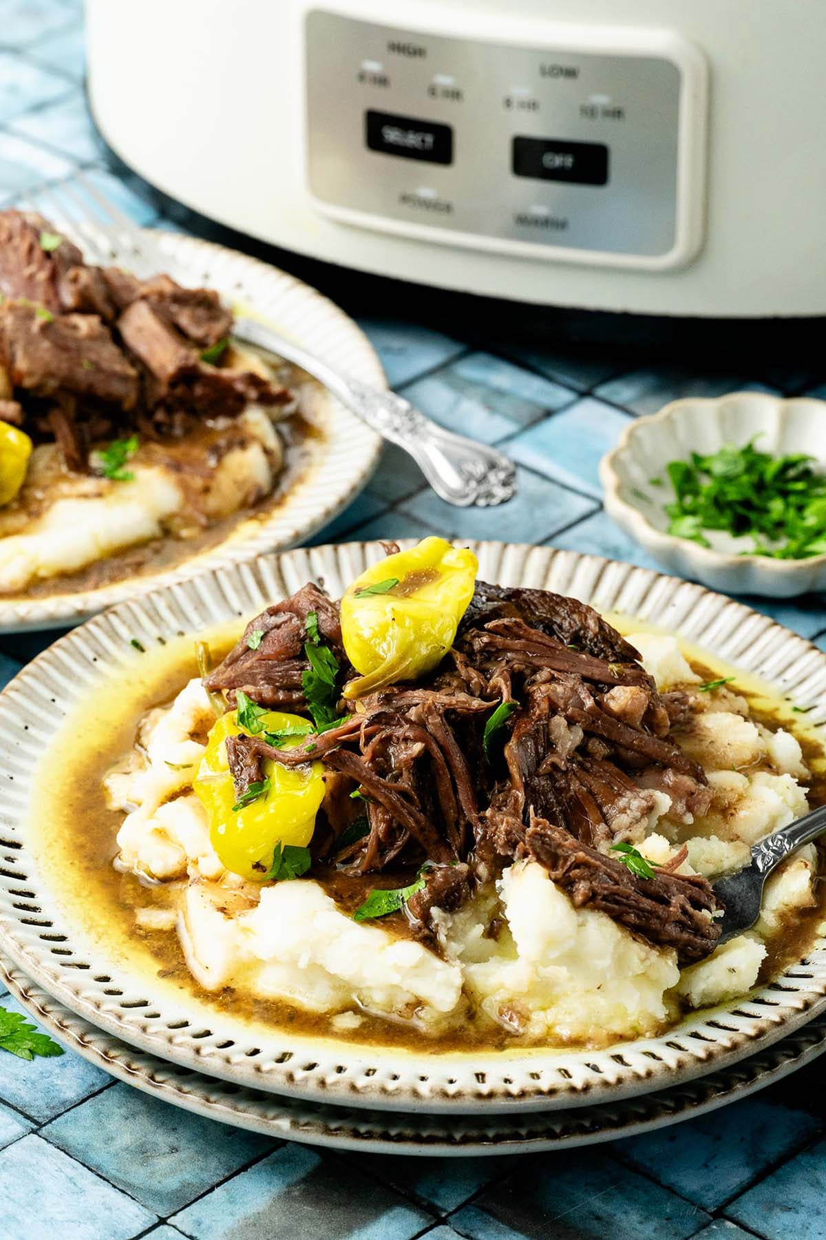 Shredded beef with peppers and gravy on mashed potatoes, served on a plate, slow cooker in background.