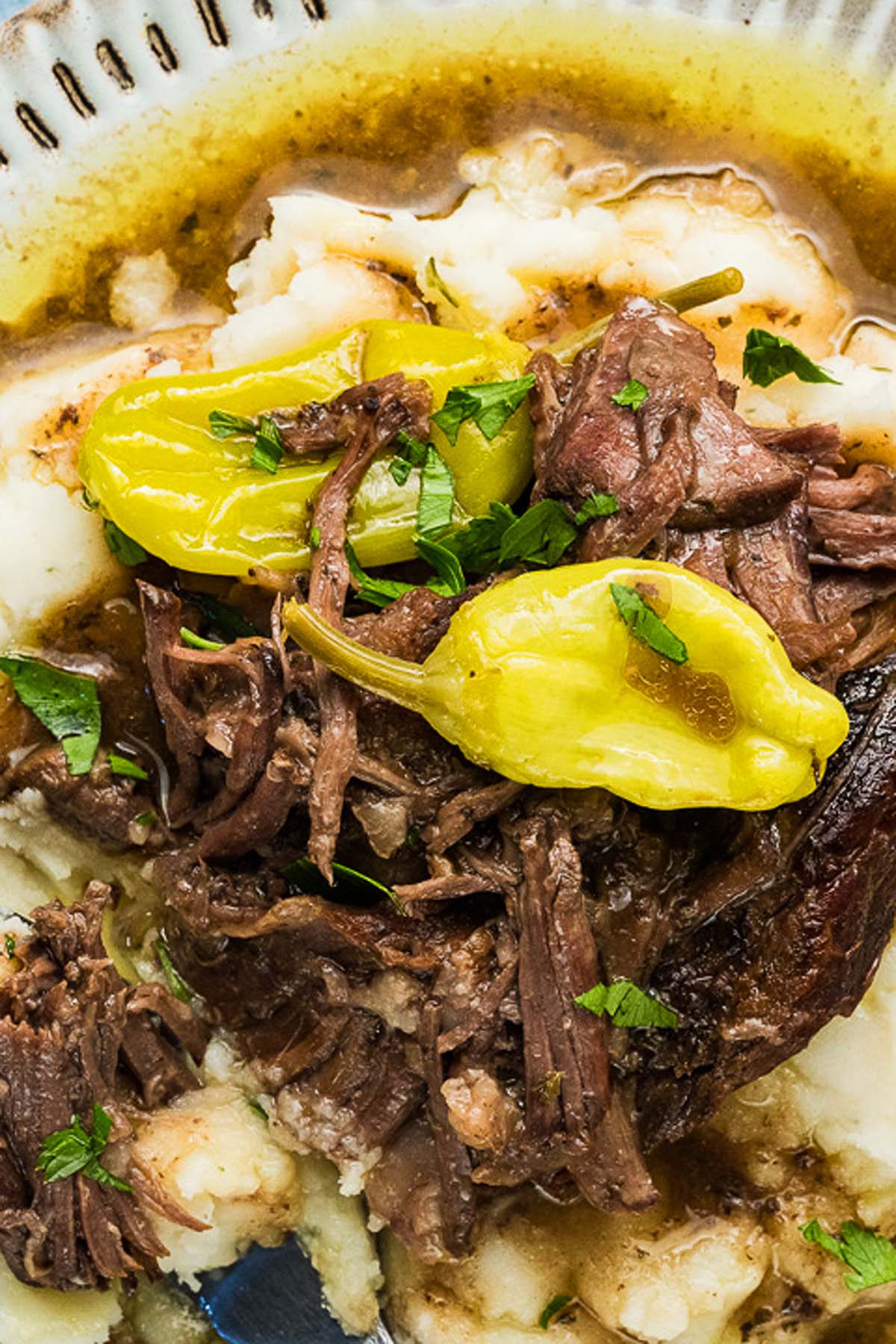 Shredded pot roast with pepperoncini peppers and gravy served over mashed potatoes on a plate.