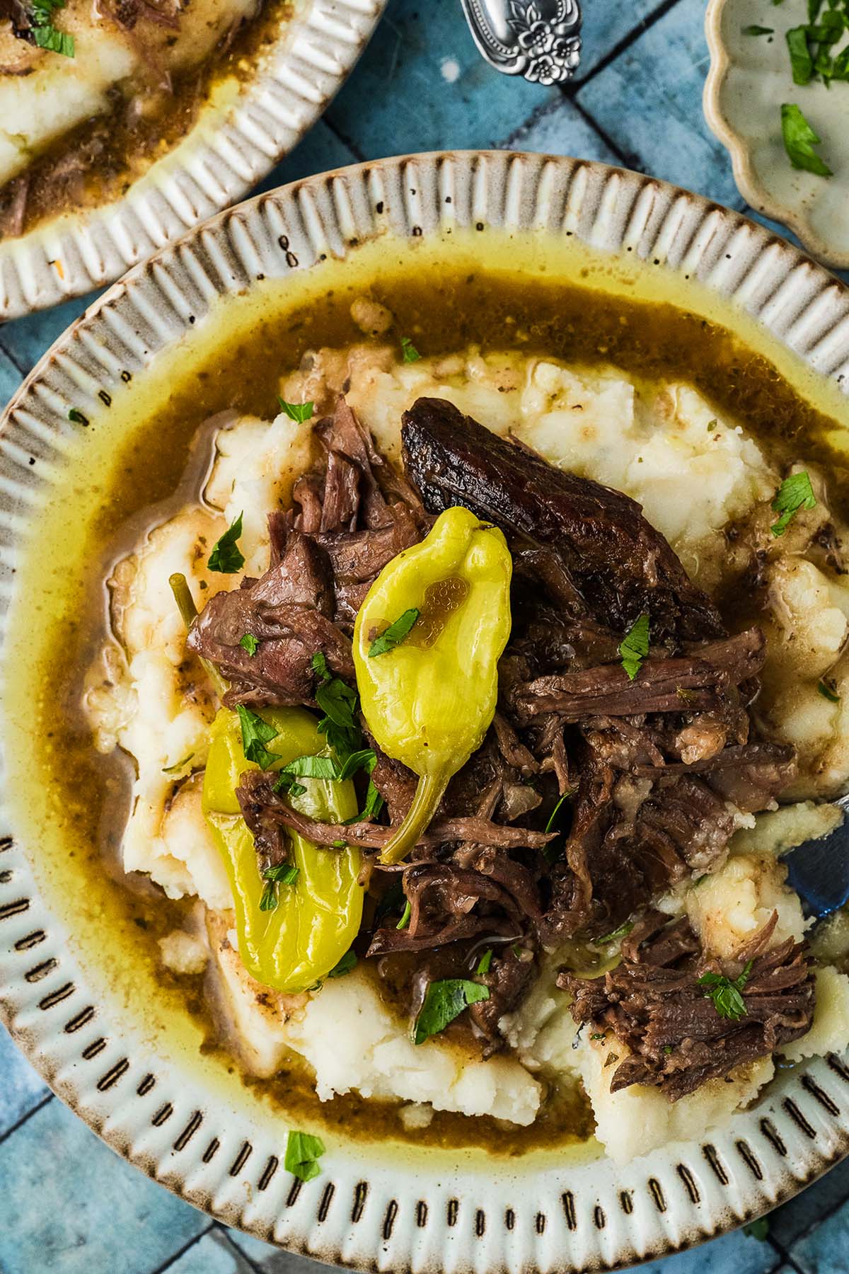 Shredded beef with pepper and gravy served over mashed potatoes on a patterned plate, garnished with herbs.