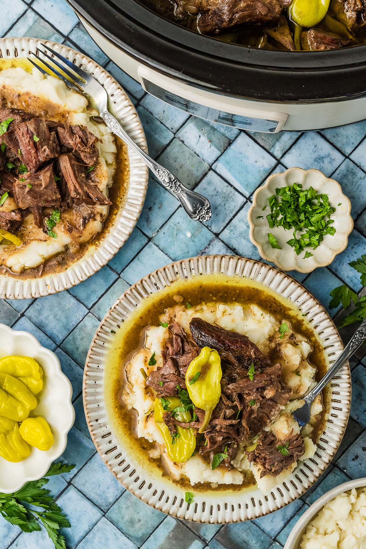 Plates of shredded beef with gravy and peppers served over mashed potatoes, surrounded by herbs and condiments.