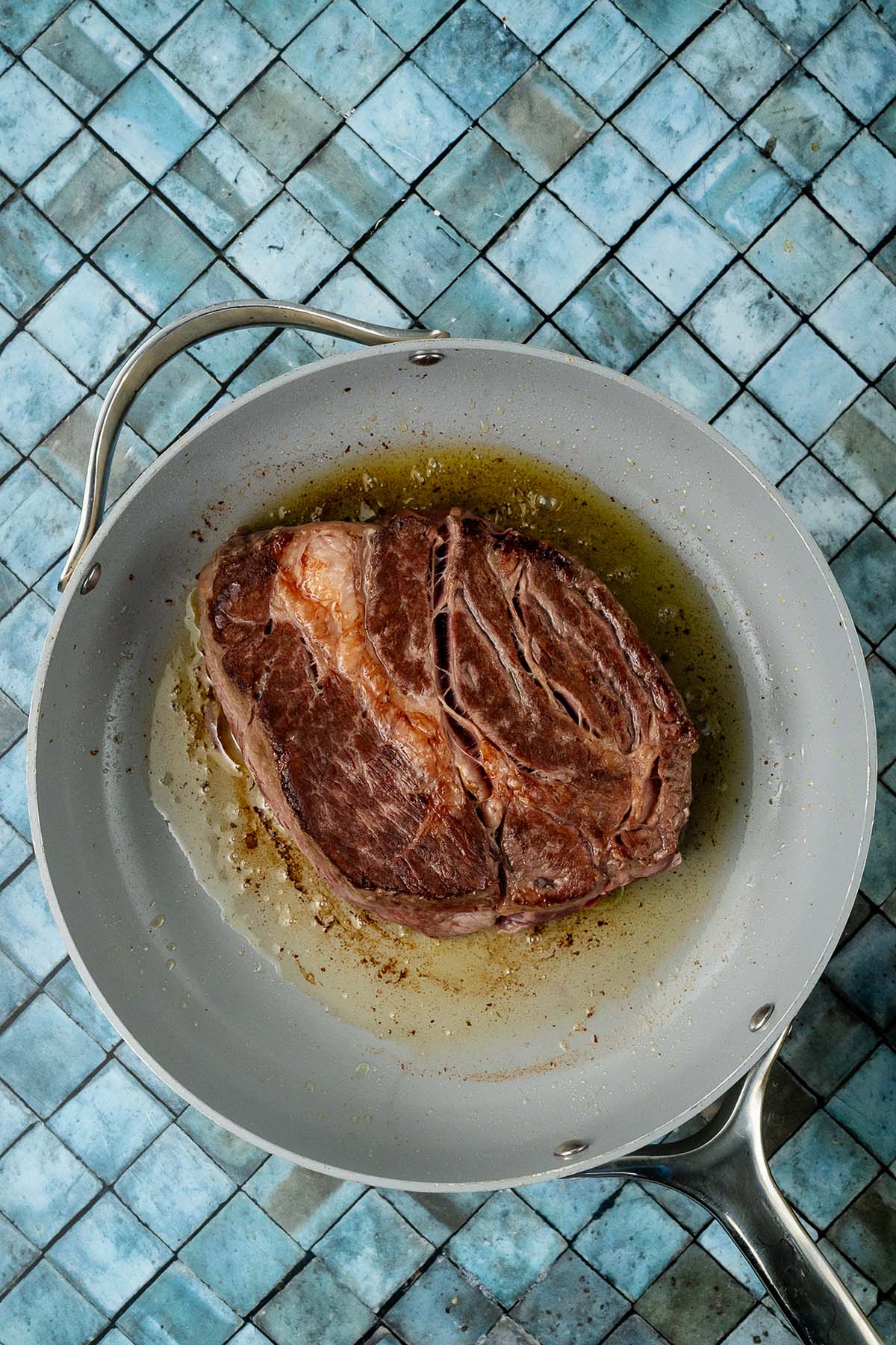 A browned roast cooking in oil in a skillet on a blue tiled surface.