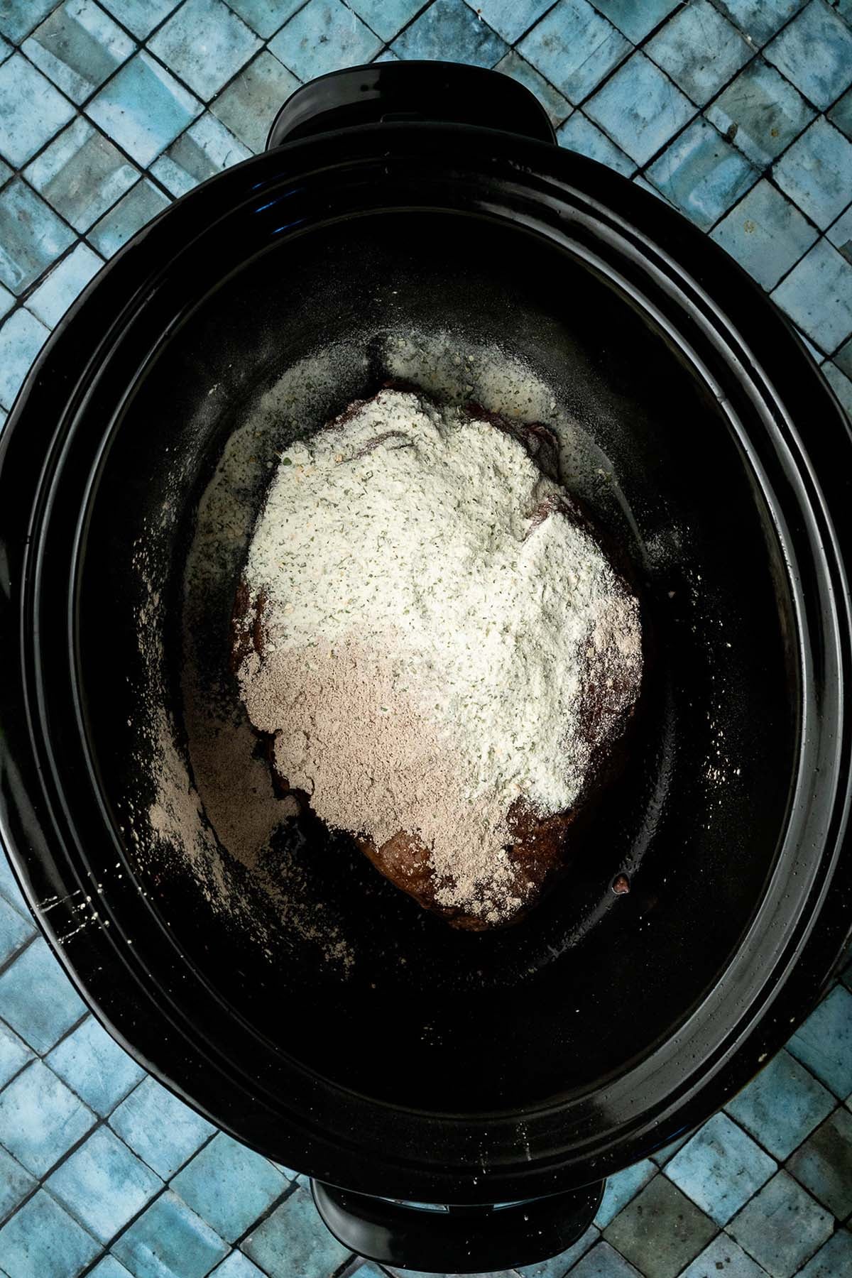 A raw beef roast topped with dry seasoning mix sits in a black slow cooker on blue tile.
