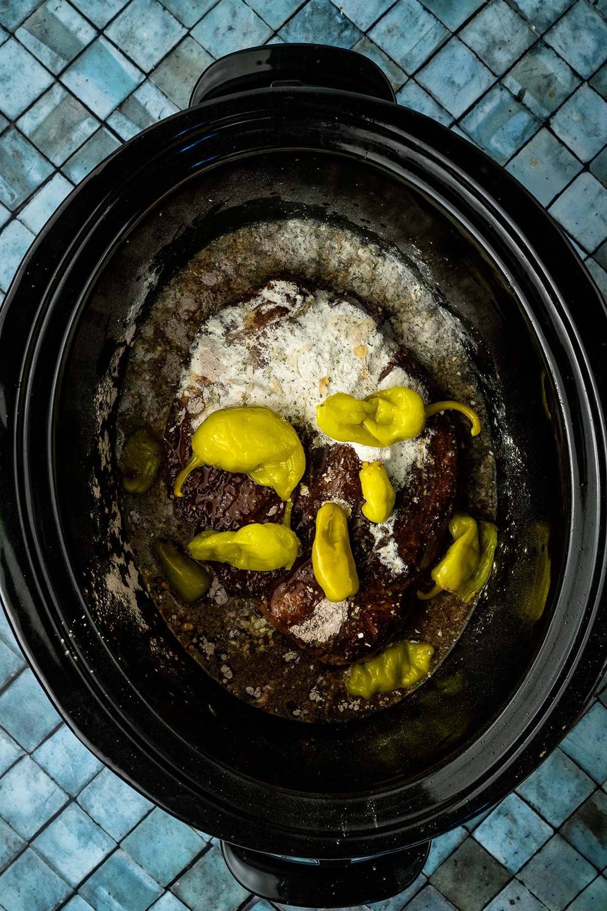 A pot roast with ranch mix and pepperoncini peppers in a black slow cooker on a blue tiled surface.