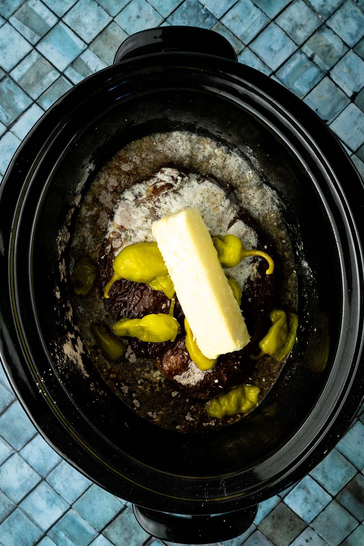 Beef roast in a slow cooker with butter, peppers, and seasoning on a blue tiled surface.
