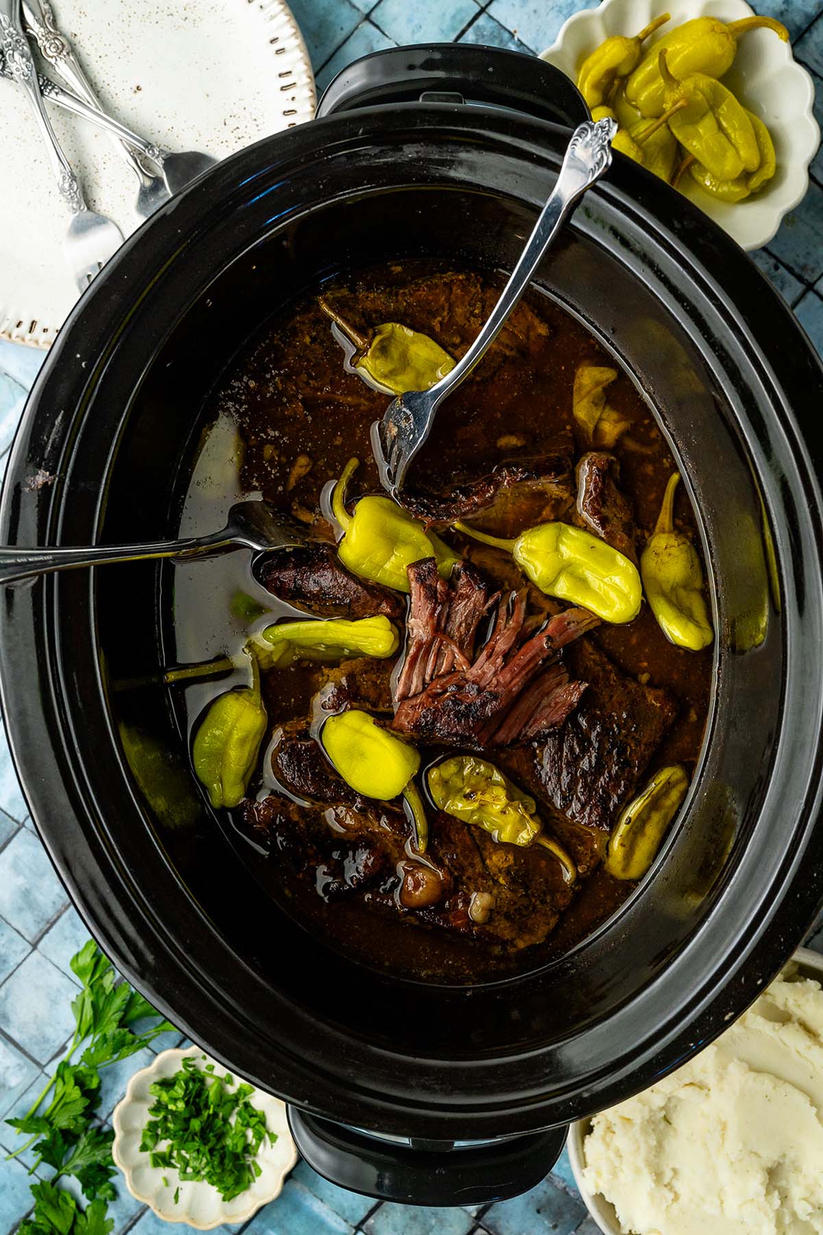 Shredded pot roast and pepperoncini in a slow cooker, served with mashed potatoes and herbs on the side.