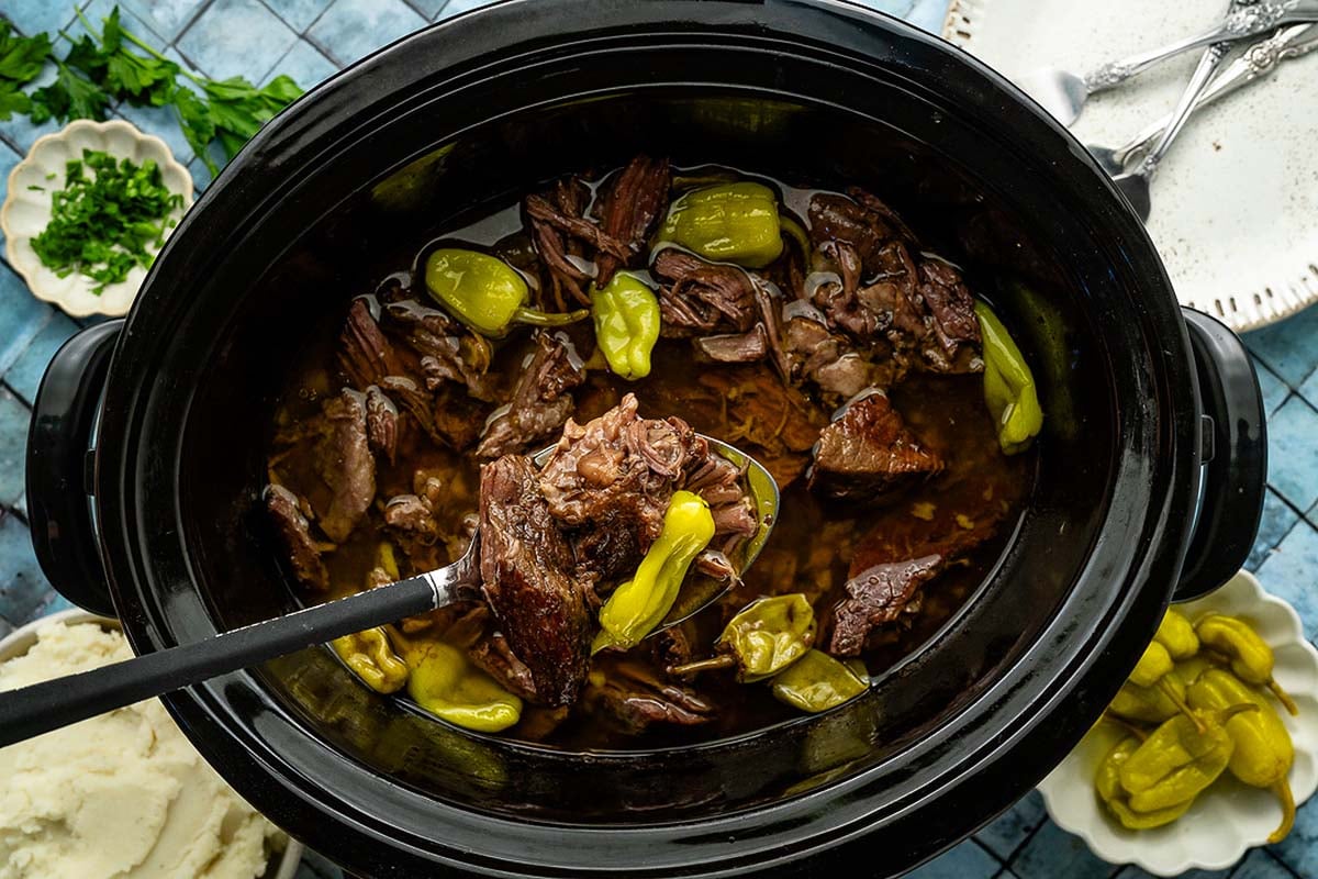 Shredded beef pot roast with pepperoncini in a slow cooker, a ladle lifting meat, with mashed potatoes on the side.