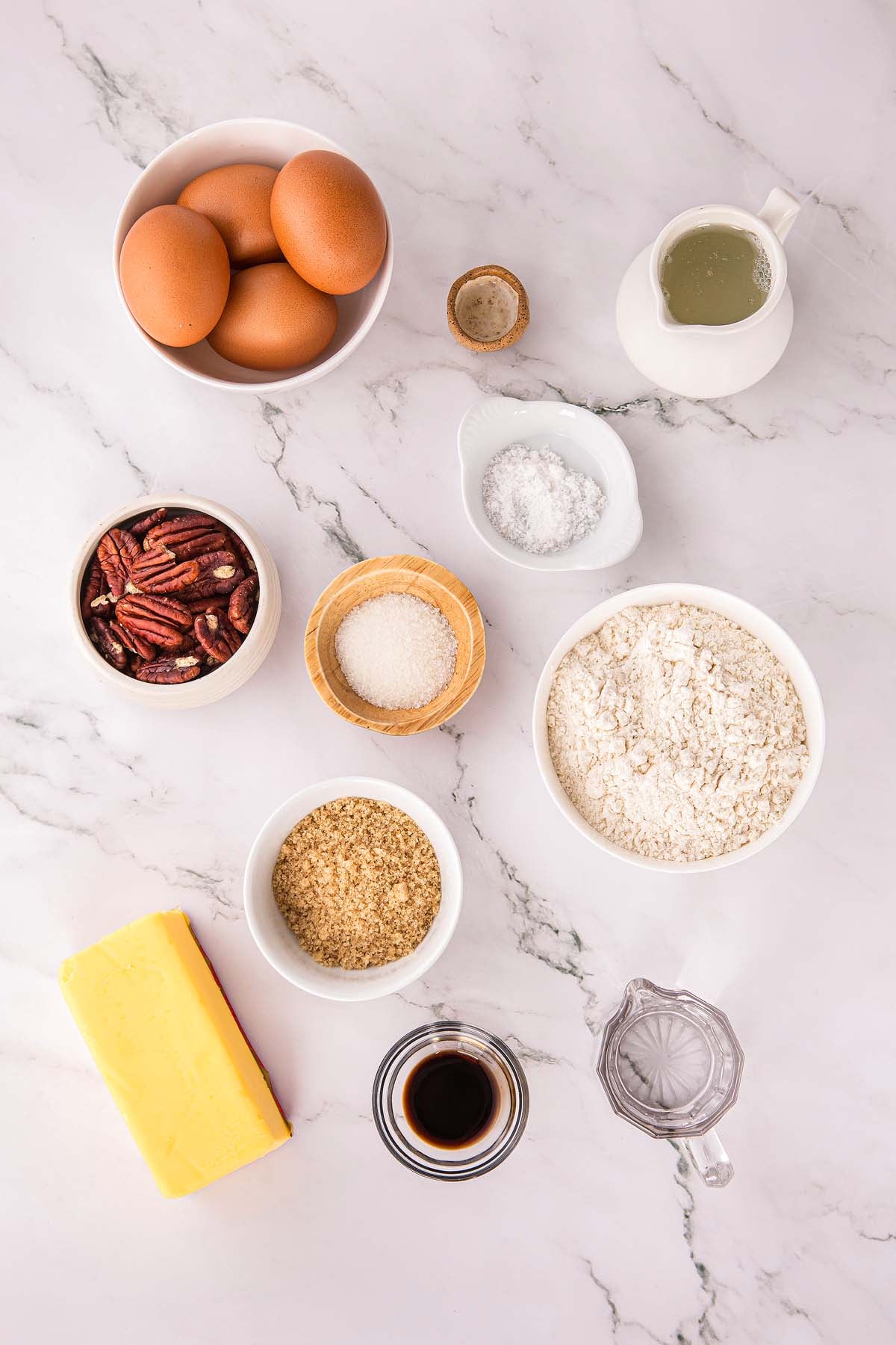 Baking ingredients on a marble surface: eggs, pecans, flour, sugar, butter, vanilla, milk, and salt.