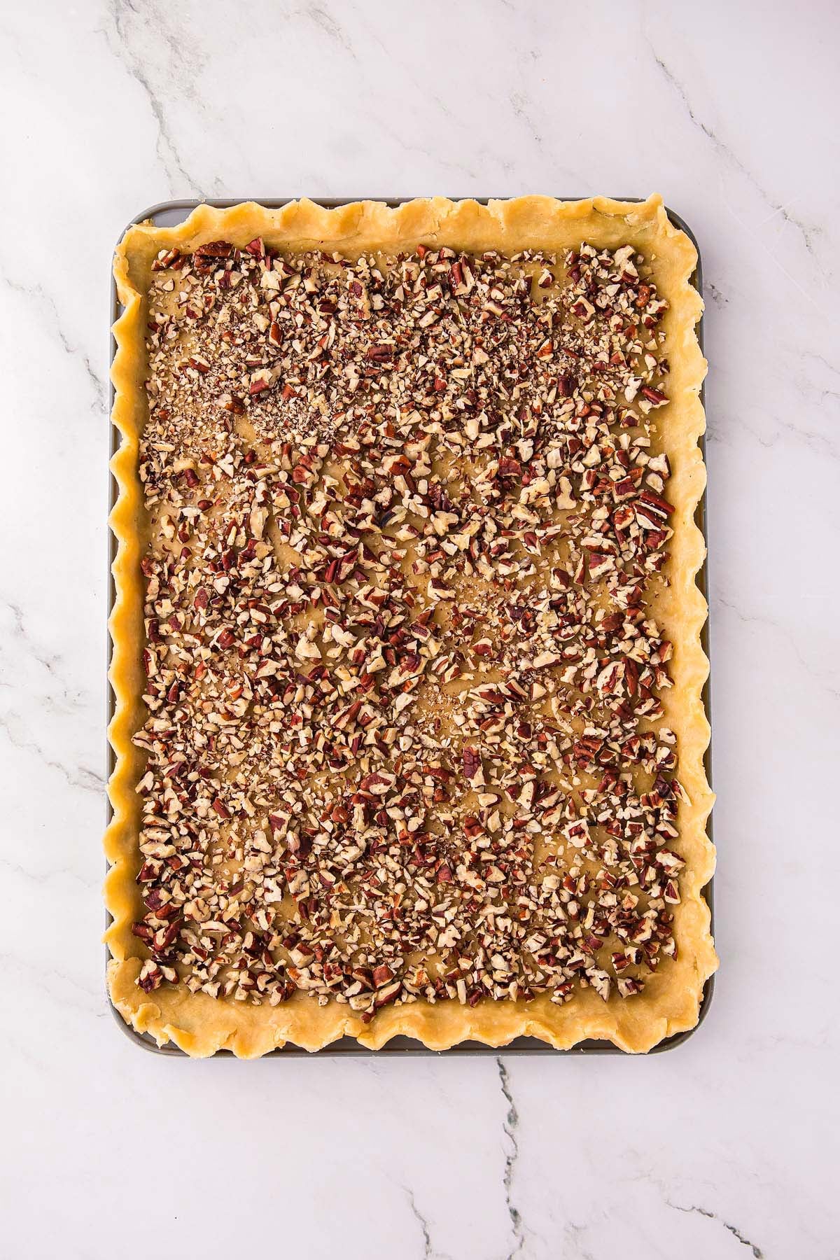 Unbaked pecan pie slab in a rectangular baking pan, topped with chopped pecans and placed on a marble surface.