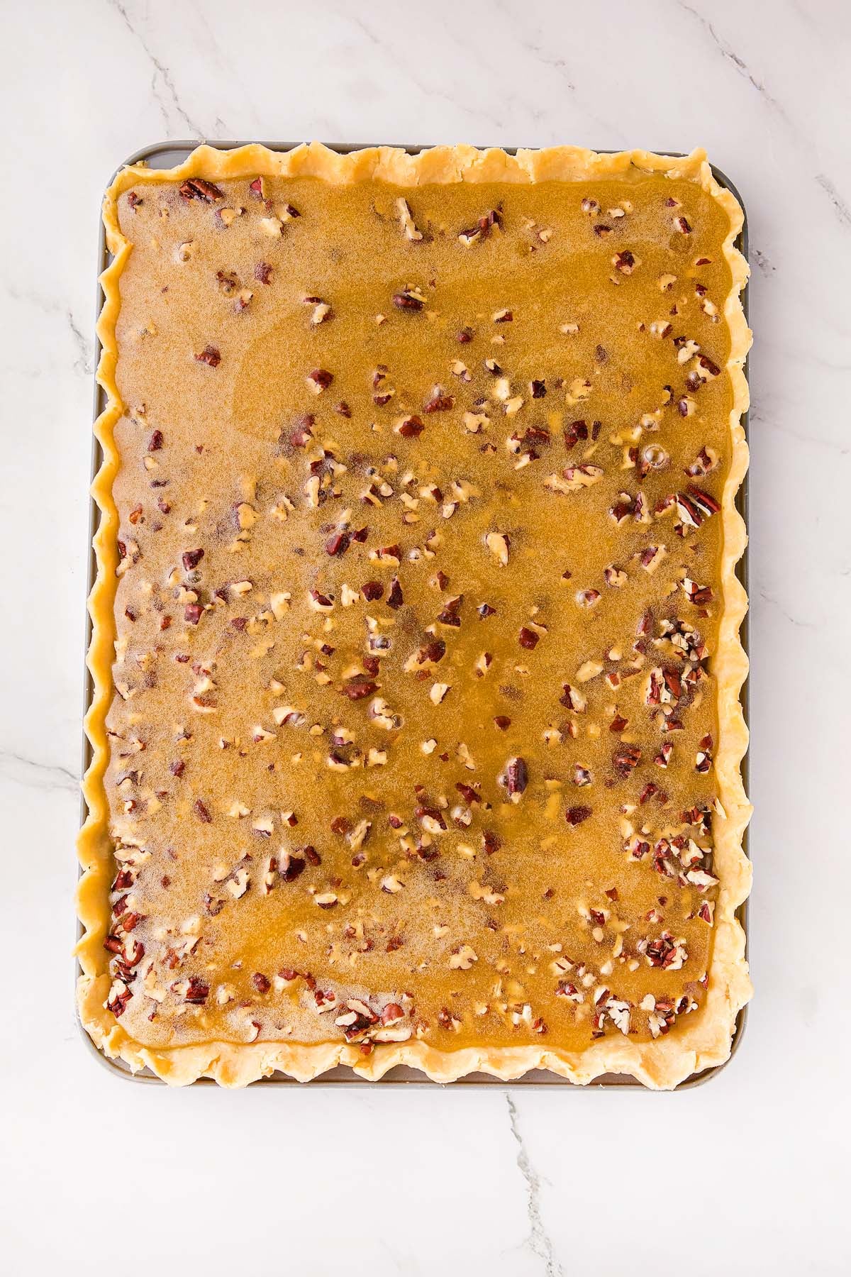 Unbaked rectangular pie crust filled with pecan mixture on a marble surface.