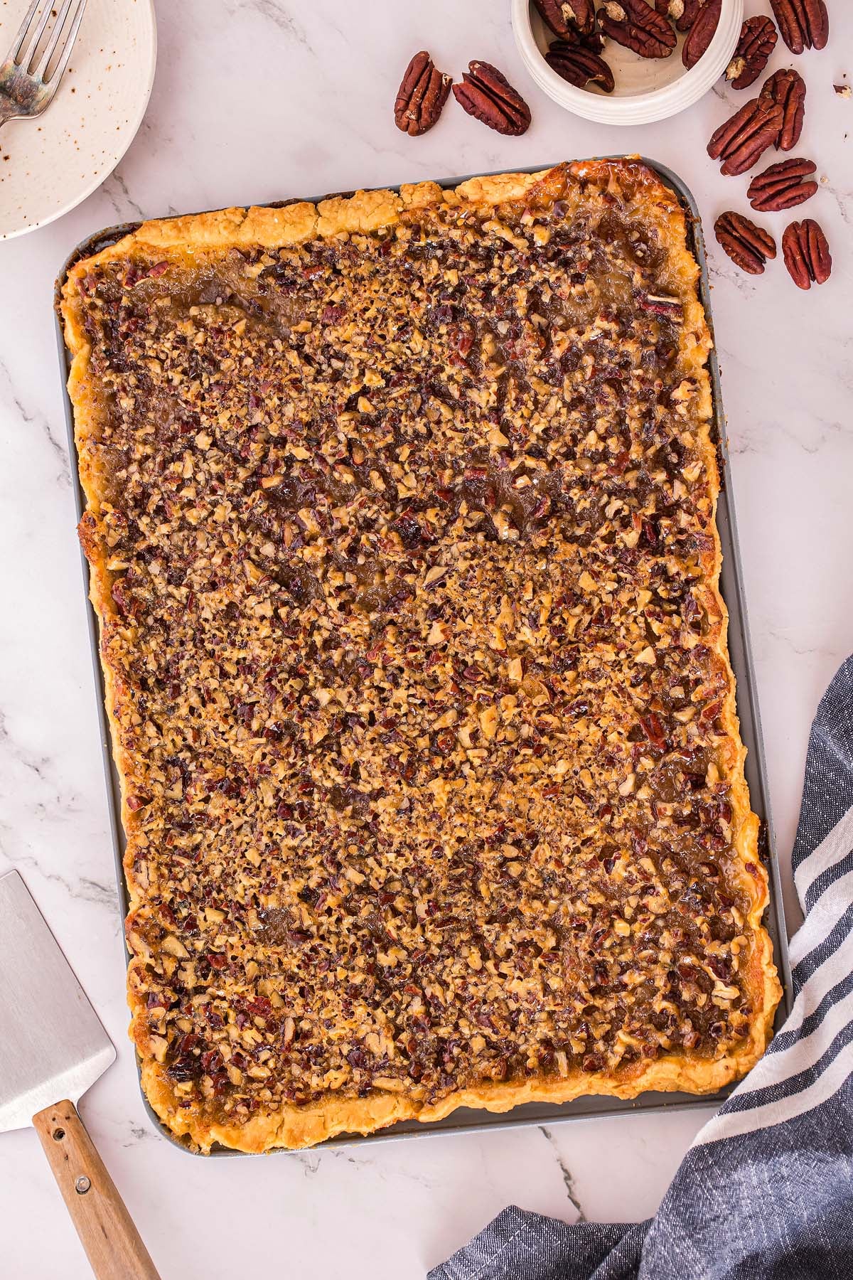 A large rectangular pecan slab pie in a baking sheet on a marble counter with pecans and utensils nearby.