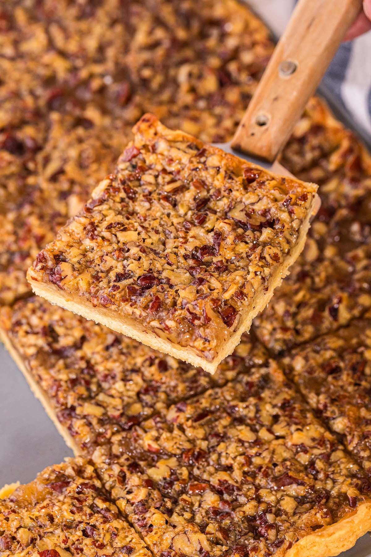 A hand lifts a pecan slab pie square from a baking tray.