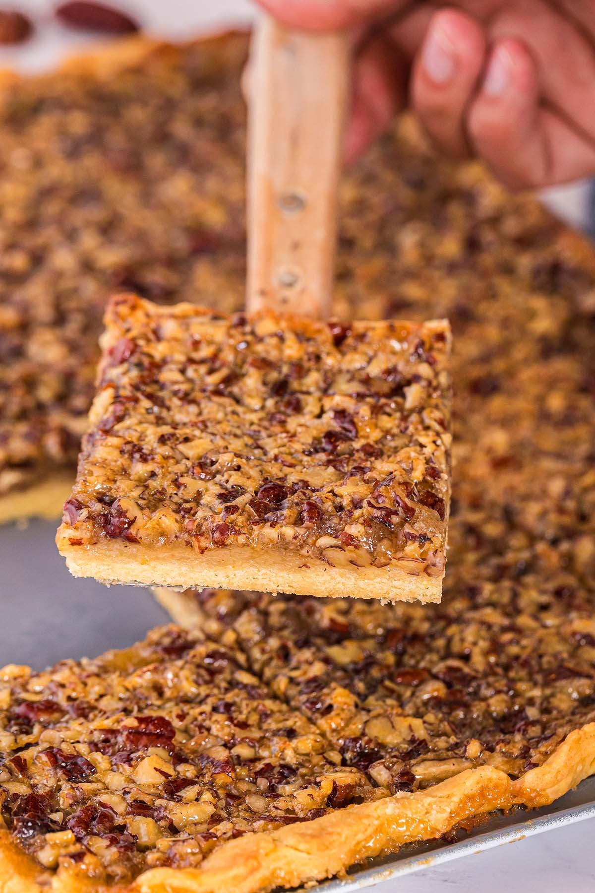 A hand lifts a square pecan slab pie slice with a spatula from a baking tray.