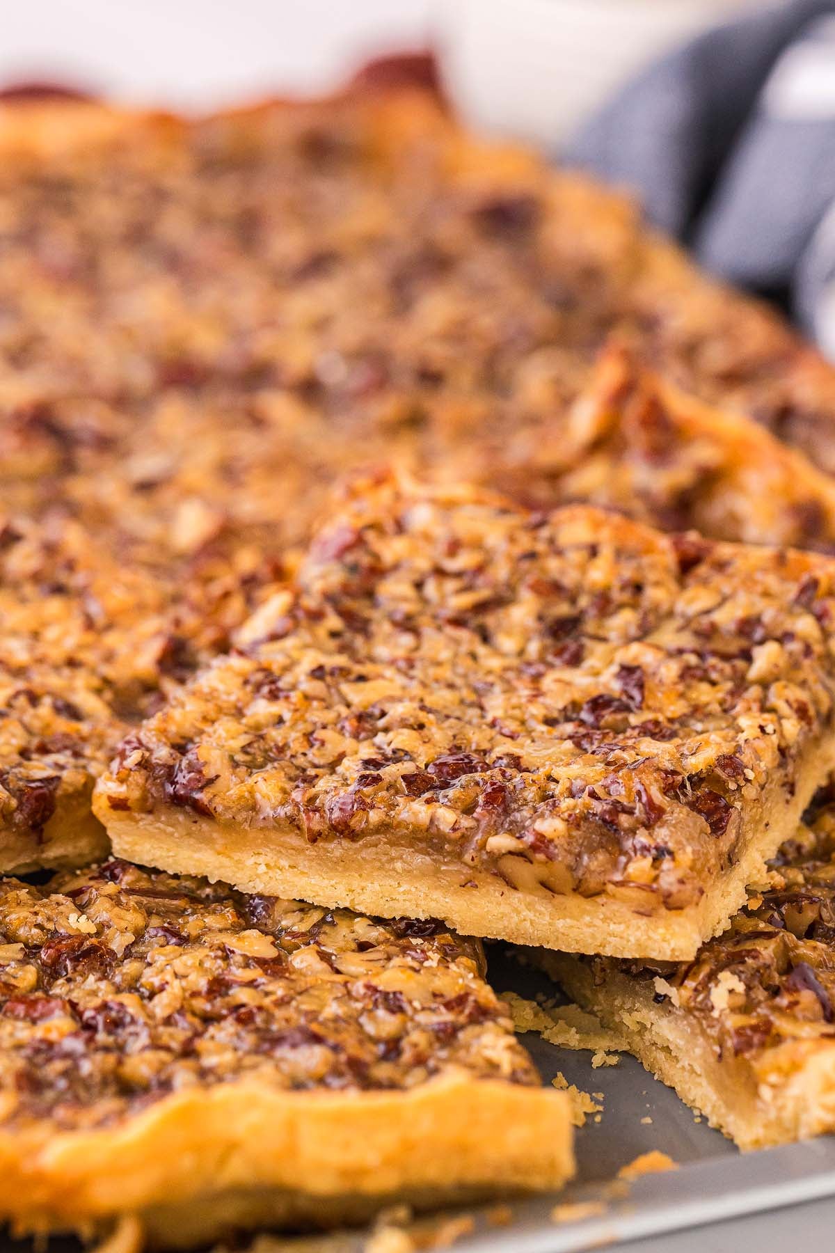 A close-up of pecan slab pie slices with a golden crust, cut into squares and stacked on a tray.