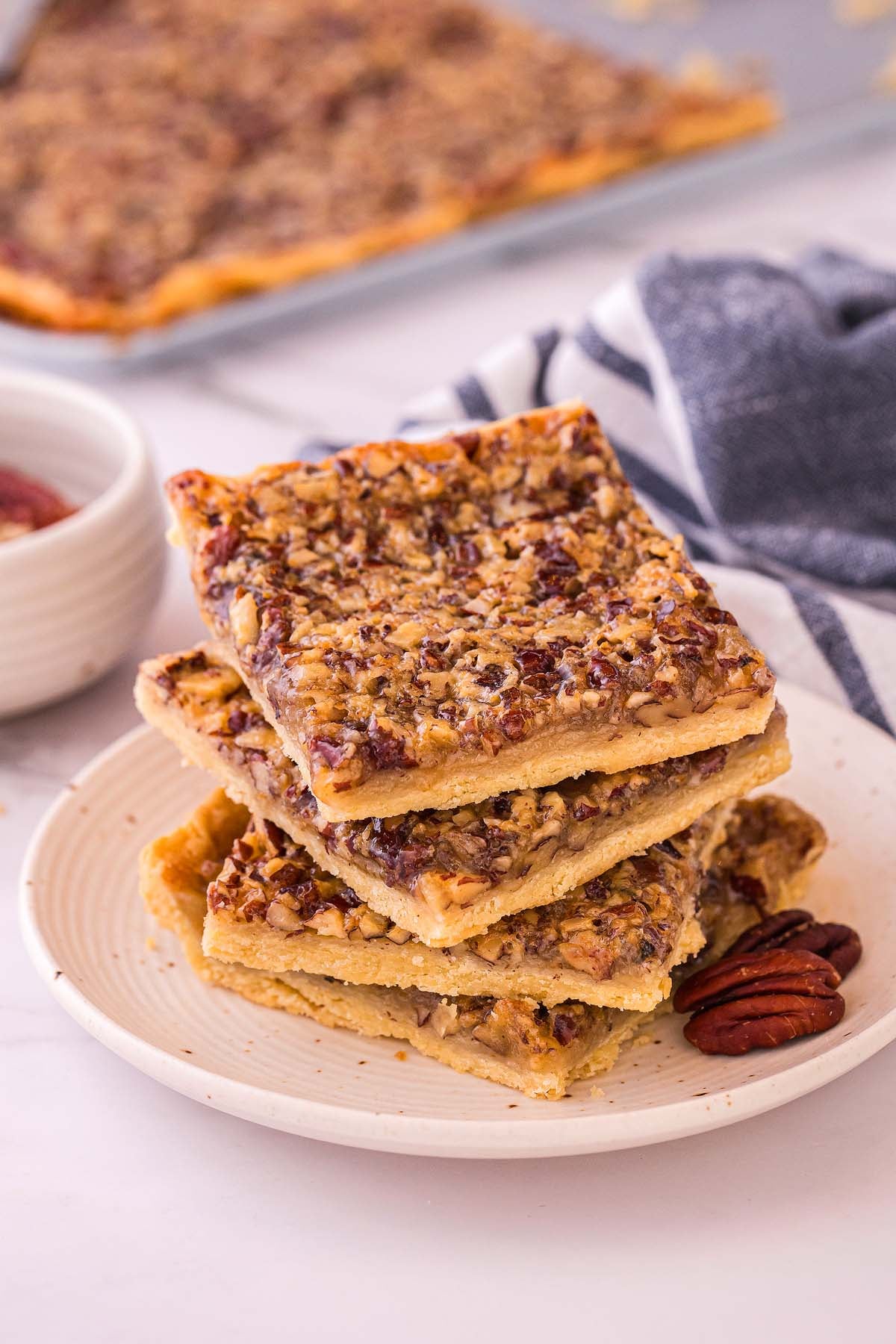 Four pecan slab pie pieces stacked on a white plate with a few whole pecans beside them, a striped towel in the background.