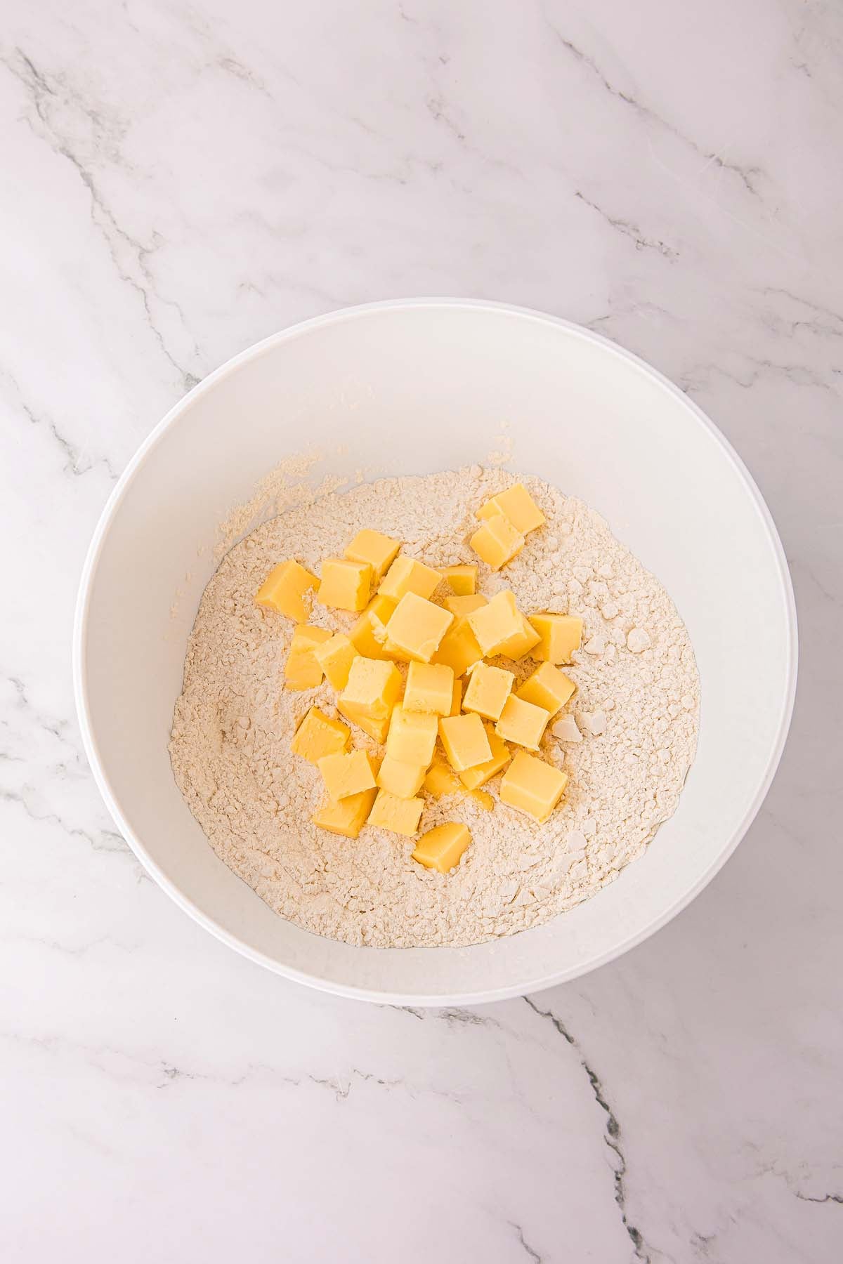 White bowl with flour and cubed butter on a marble surface, viewed from above.