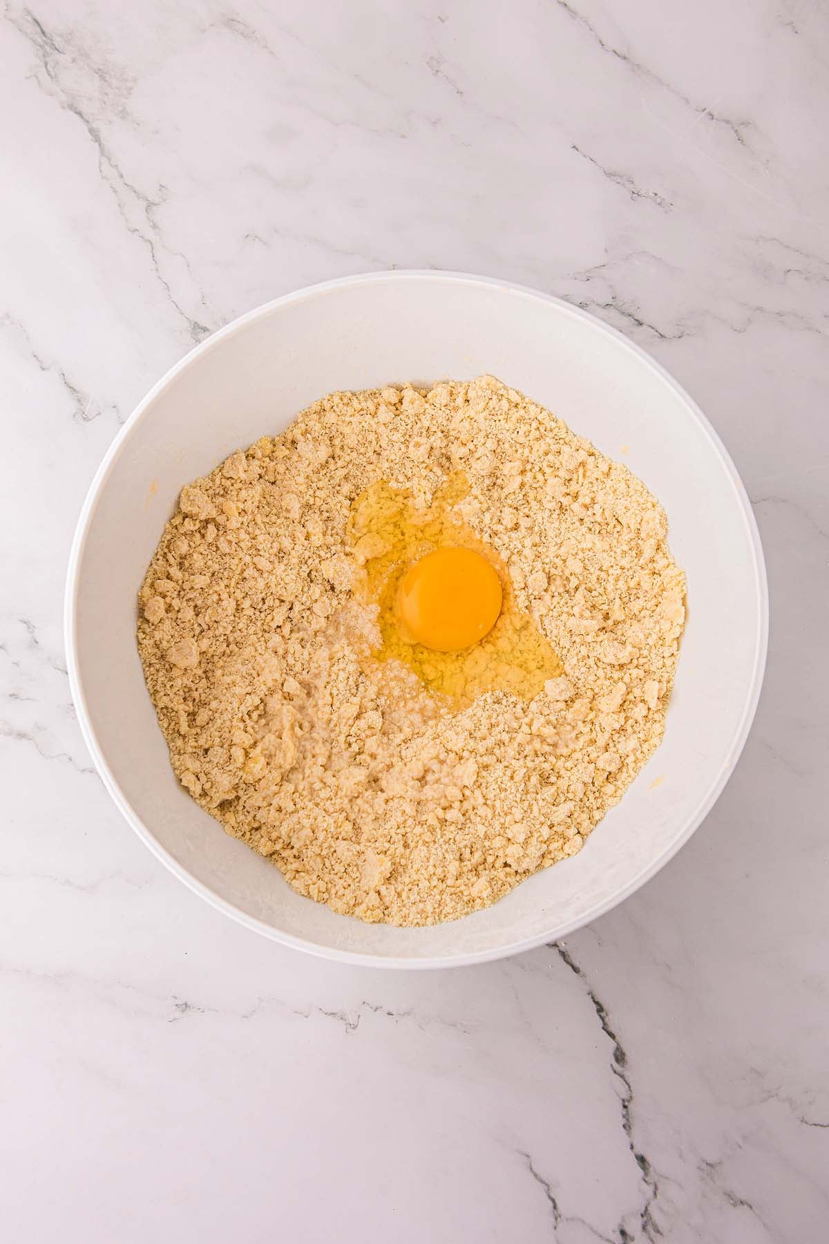 A cracked egg sits atop flour mixture in a white mixing bowl on a marble countertop.