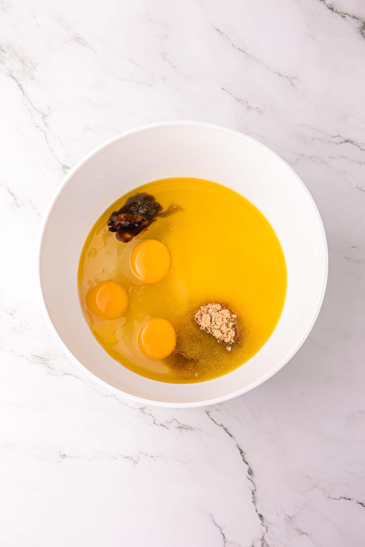 Three raw eggs, brown sugar, and vanilla in a white bowl on a marble countertop.