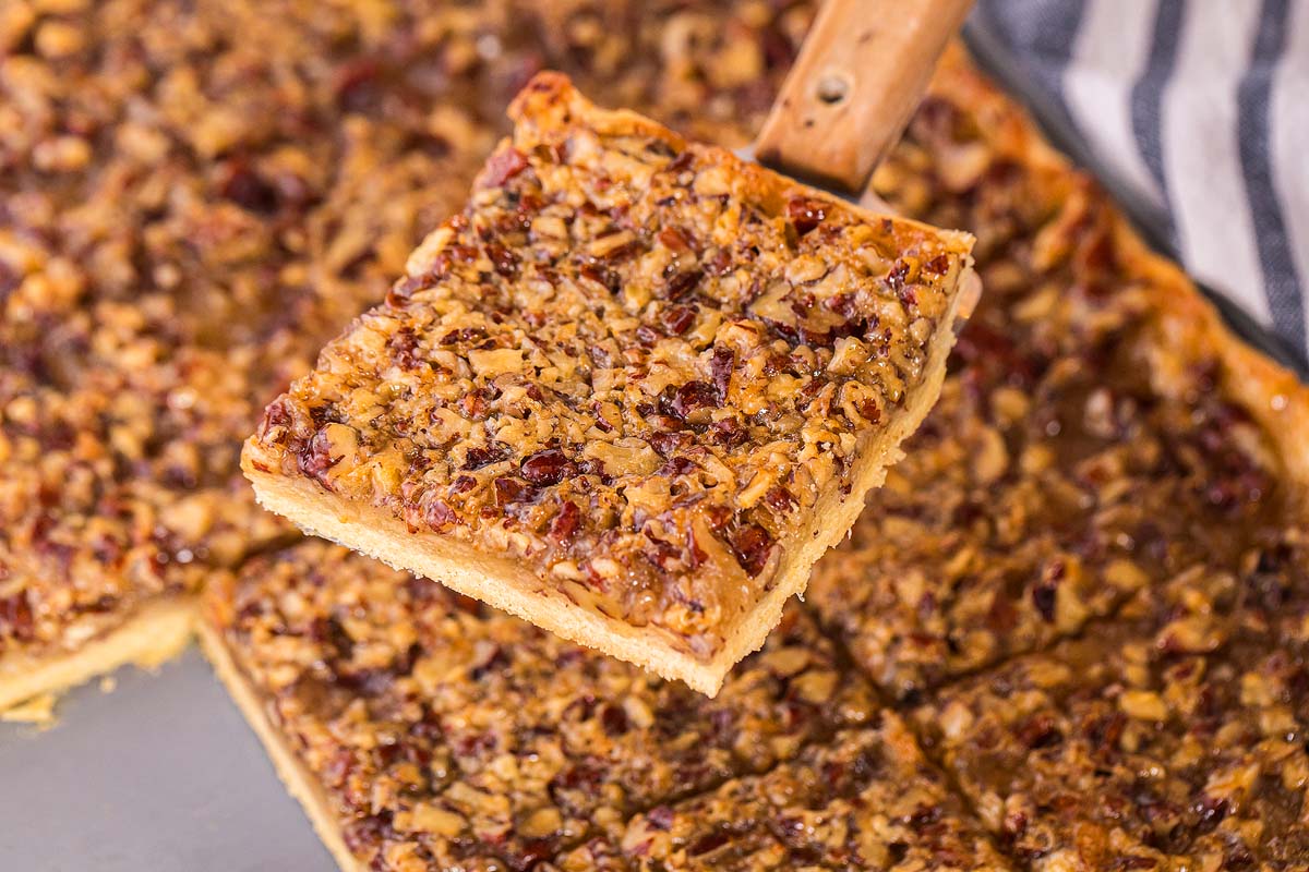 A close-up of a pecan slab pie piece being lifted from a pan with a metal spatula.