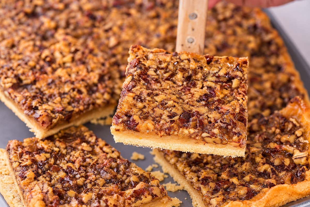 A spatula lifts a square pecan slab pie piece from a baking tray.
