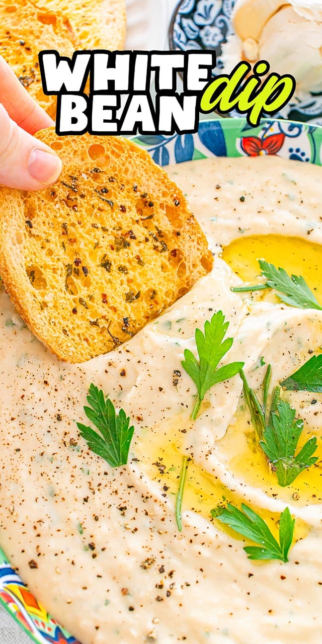 A hand dips toasted bread into creamy white bean dip, garnished with parsley, pepper, and olive oil.
