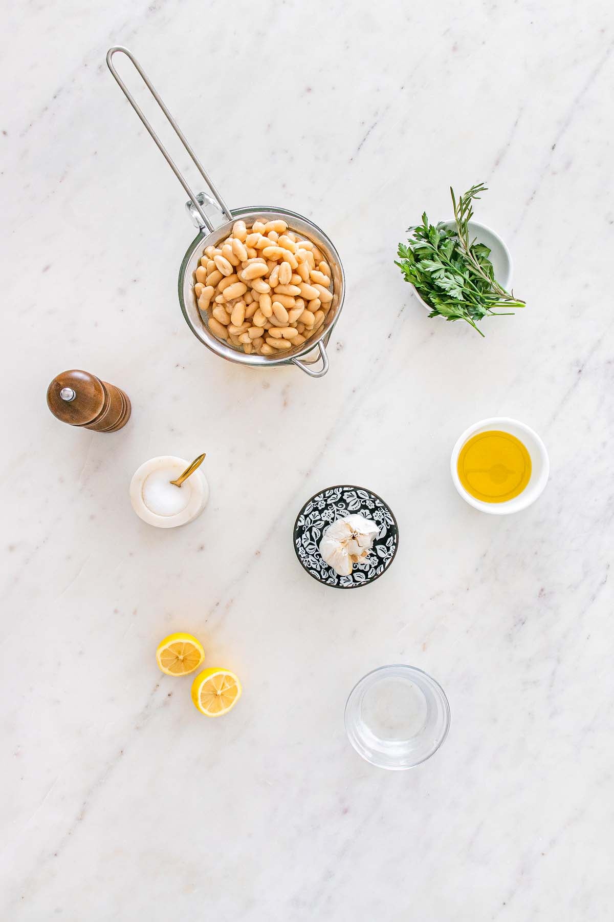 Ingredients on a marble counter: white beans, oil, herbs, garlic, lemon, salt, pepper, and water.