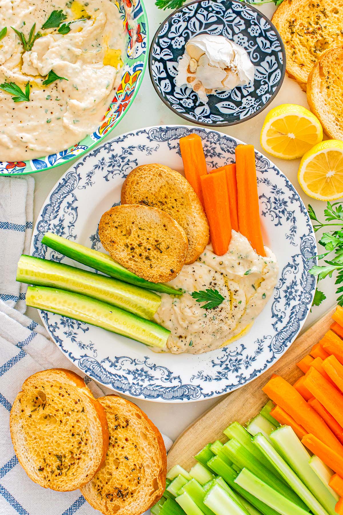 Plate with white bean dip, toasted bread, carrot and cucumber sticks, and lemon halves on the side.