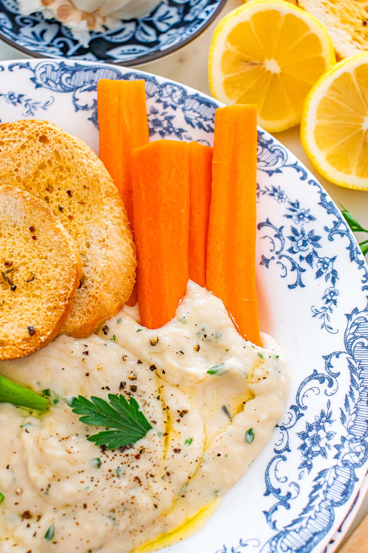 Three carrot sticks, bread slices, and creamy Tuscan cannellini dip garnished with parsley on a blue and white plate, lemon nearby.