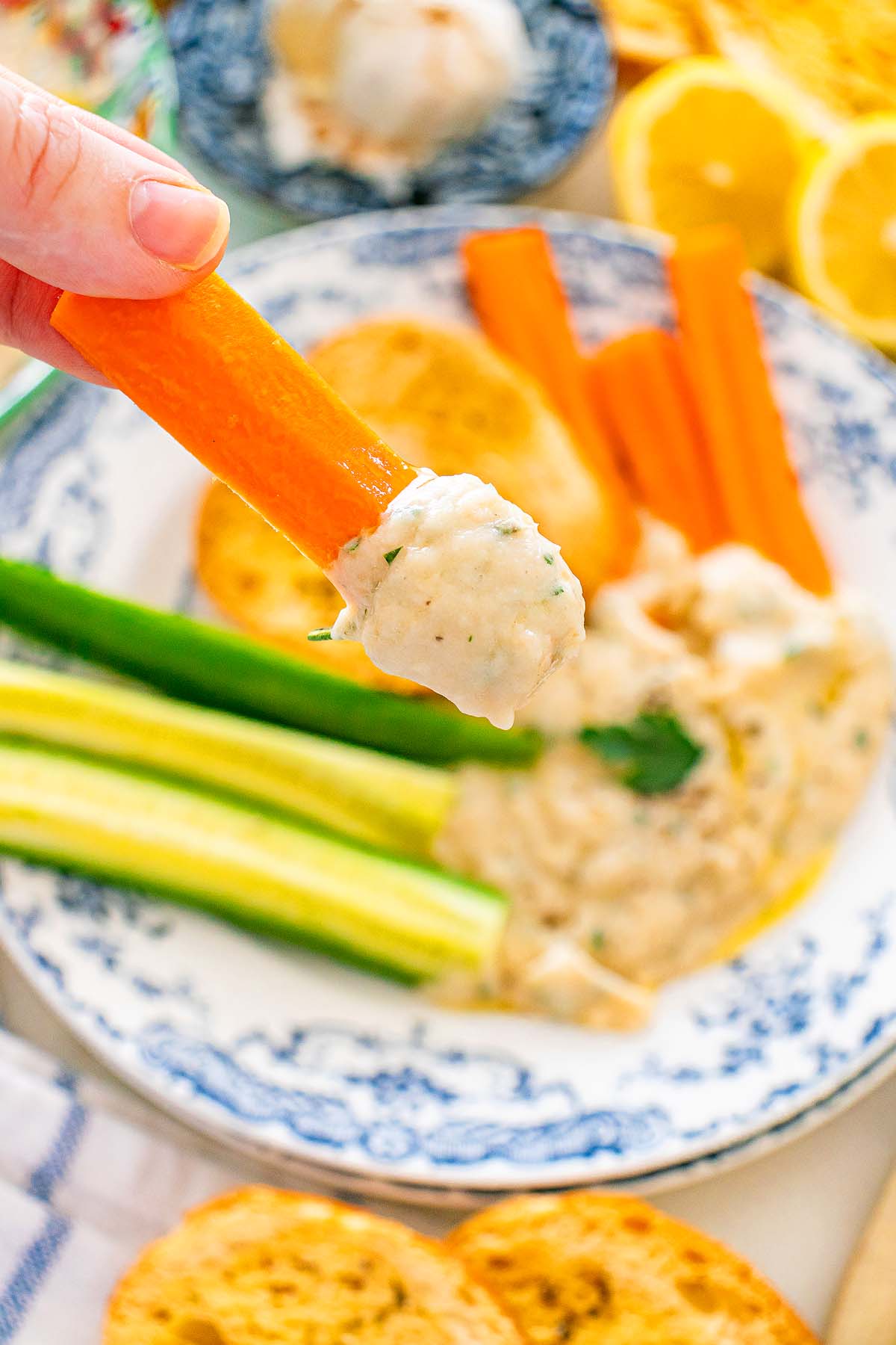 A hand dips a carrot stick into a creamy dip, with more veggies and dip on a plate in the background.